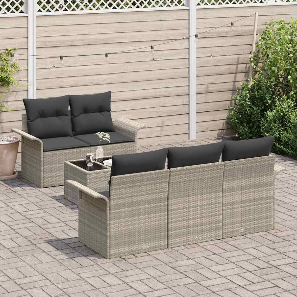 Outdoor-Terrassenset: zwei Sofas und ein kleiner Tisch. Graue Korbmöbel mit schwarzen Kissen. Hintergrund ist ein Holzzaun und Pflanzen.