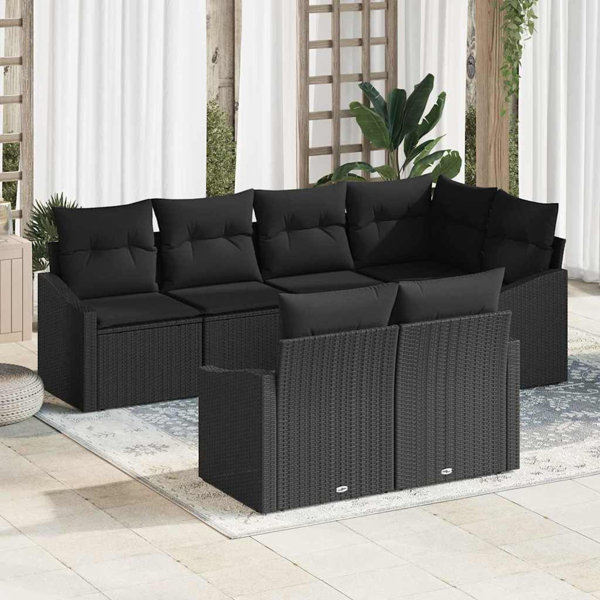 Ein schwarzes Outdoor-Ecksofa aus Korbgeflecht mit schwarzen Kissen steht auf einer Terrasse. Weiße Vorhänge sind im Hintergrund. Ein Teppich liegt unter dem Sofa.