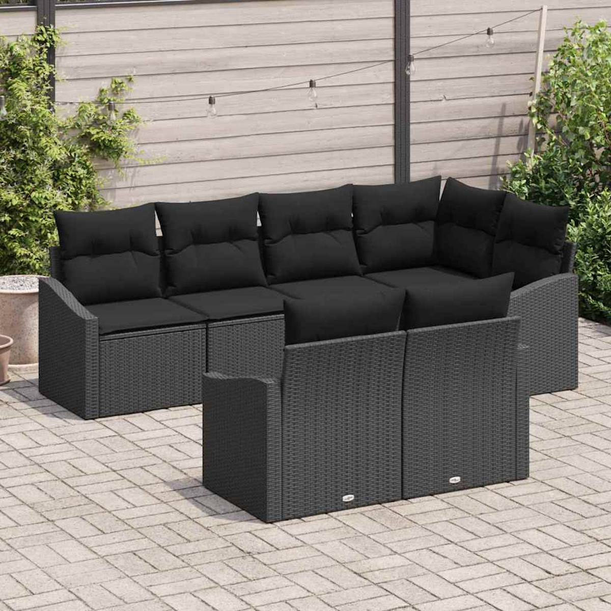 Outdoor-Ecksofa mit schwarzen Kissen auf einer Steinterrasse. Der Hintergrund umfasst einen Zaun, Grün und Lichterketten.