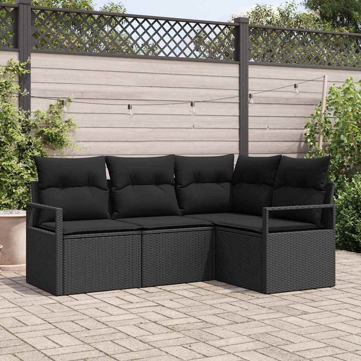Schwarzes Rattan-Ecksofa mit schwarzen Kissen auf einer Ziegelterrasse. Der Hintergrund umfasst einen Zaun mit Lichtern und Grün.