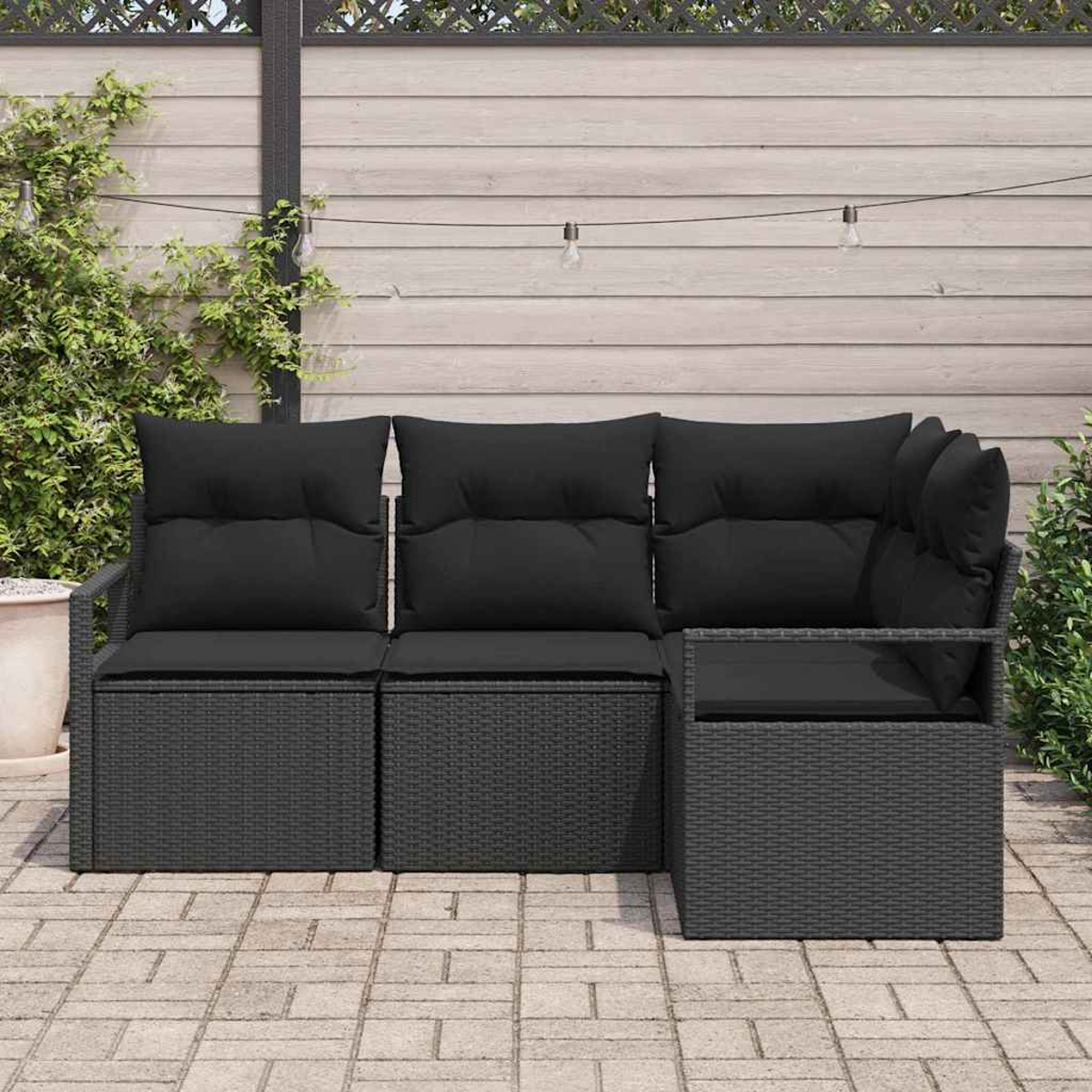 Schwarzes Outdoor-Ecksofa aus Korbgeflecht mit schwarzen Kissen auf einer Backstein-Terrasse. Im Hintergrund ein Holzzaun und Pflanzen.