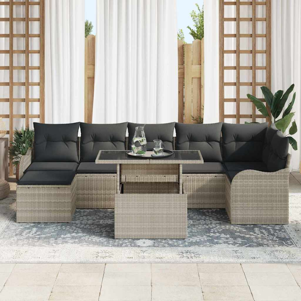 Outdoor-Rattan-Sofagarnitur mit dunkelgrauen Kissen. Davor steht ein Glastisch mit Getränken. Weiße Vorhänge und Holzzaun im Hintergrund.