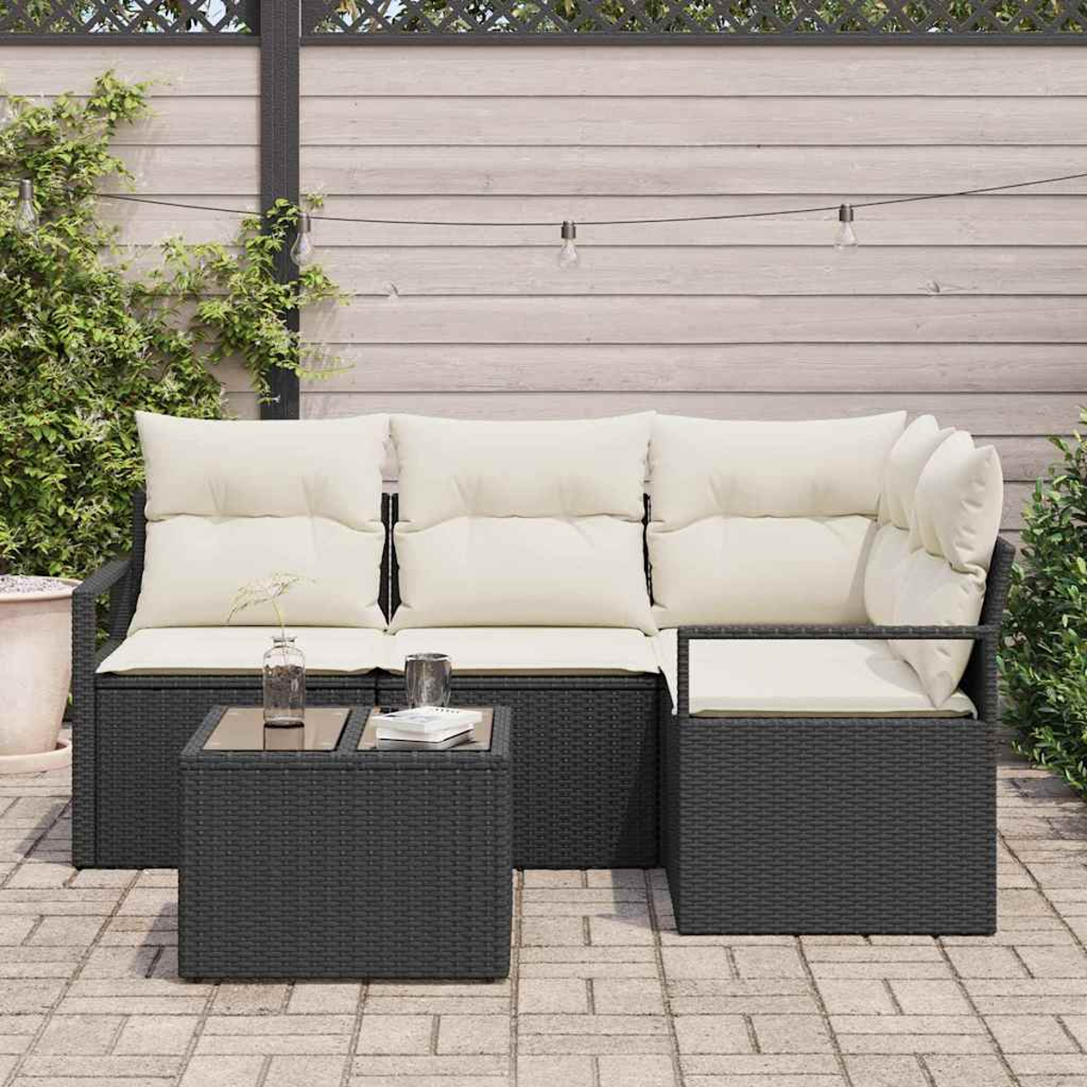 Schwarzes Outdoor-Korbsitzgarnitur mit cremefarbenen Kissen und einem kleinen Tisch. Im Hintergrund ein Holzzaun und Lichterketten.