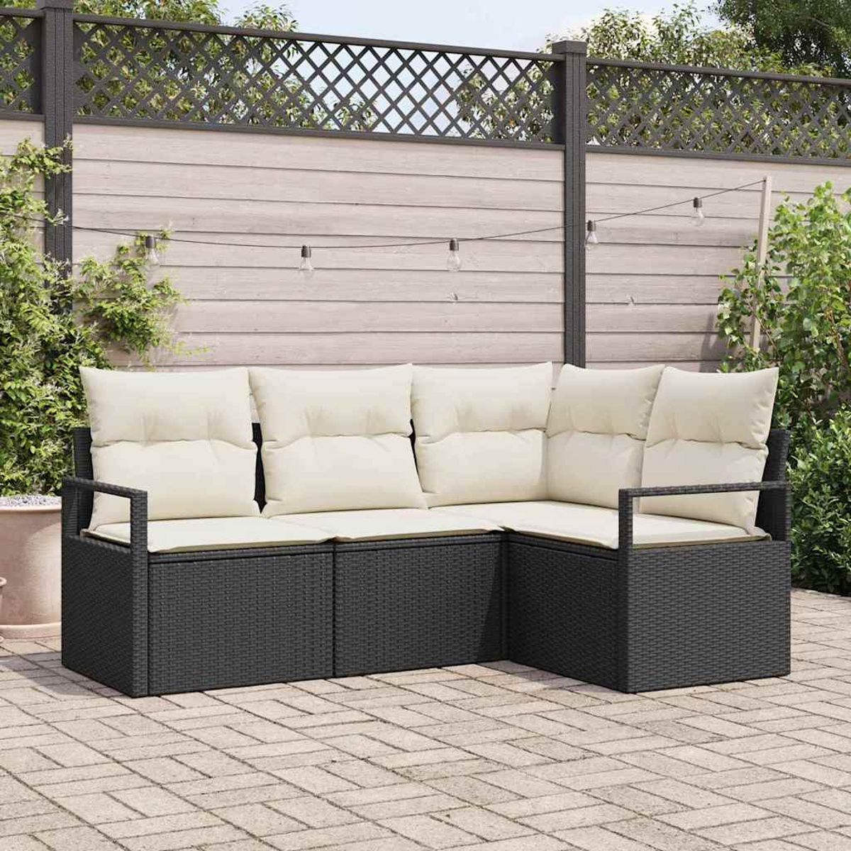 Ein schwarzes Korbsessel-Ecksofa mit cremefarbenen Kissen steht auf einer Backstein-Terrasse. Im Hintergrund ein Holzzaun mit dekorativem Gitter.