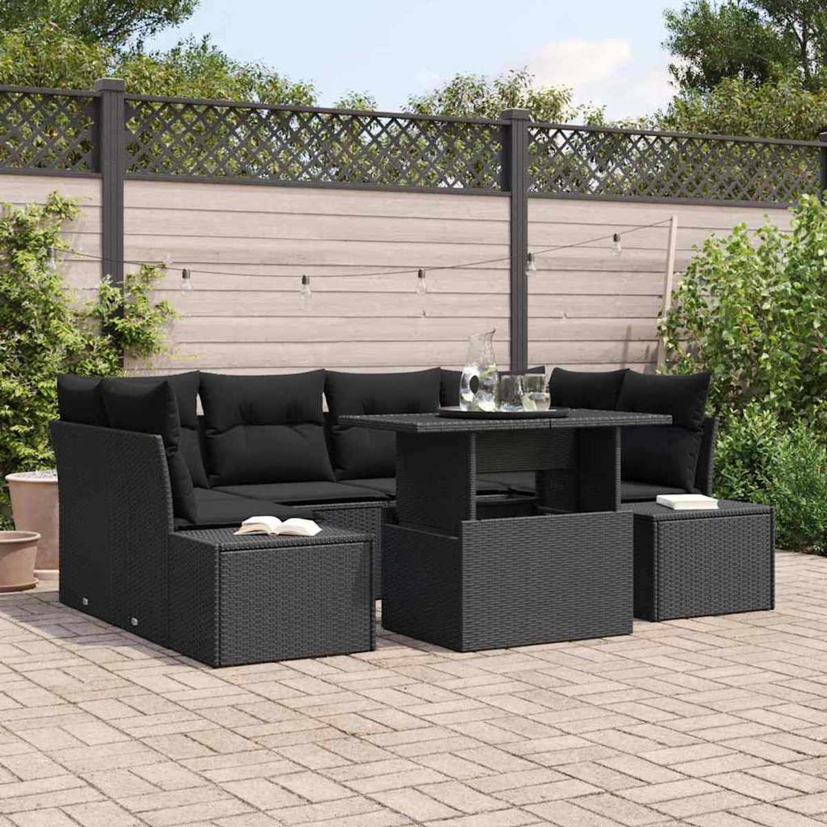 Schwarzes Gartenmöbel-Set aus Korbgeflecht mit Tisch, Sofa und Ottomanen auf einer Ziegelterrasse, mit Zaun und Pflanzen im Hintergrund.