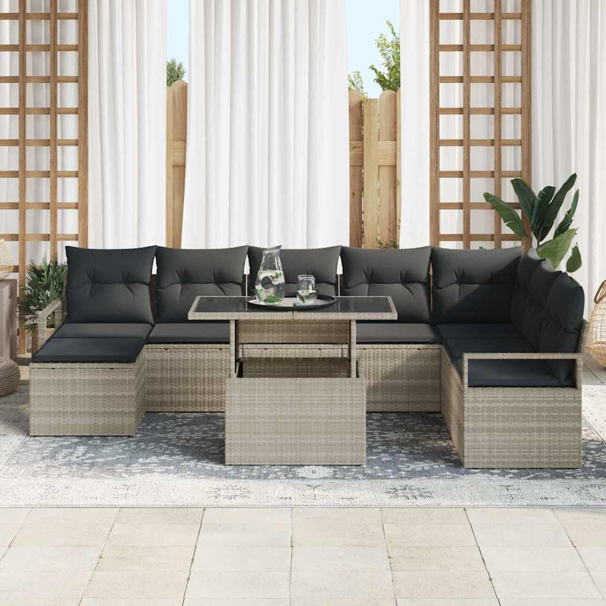 Outdoor-Ecksofa mit schwarzen Kissen. Ein Tisch mit Glasplatte trägt Wasserkrug und Gläser. Weiße Vorhänge und Holzzaun im Hintergrund.