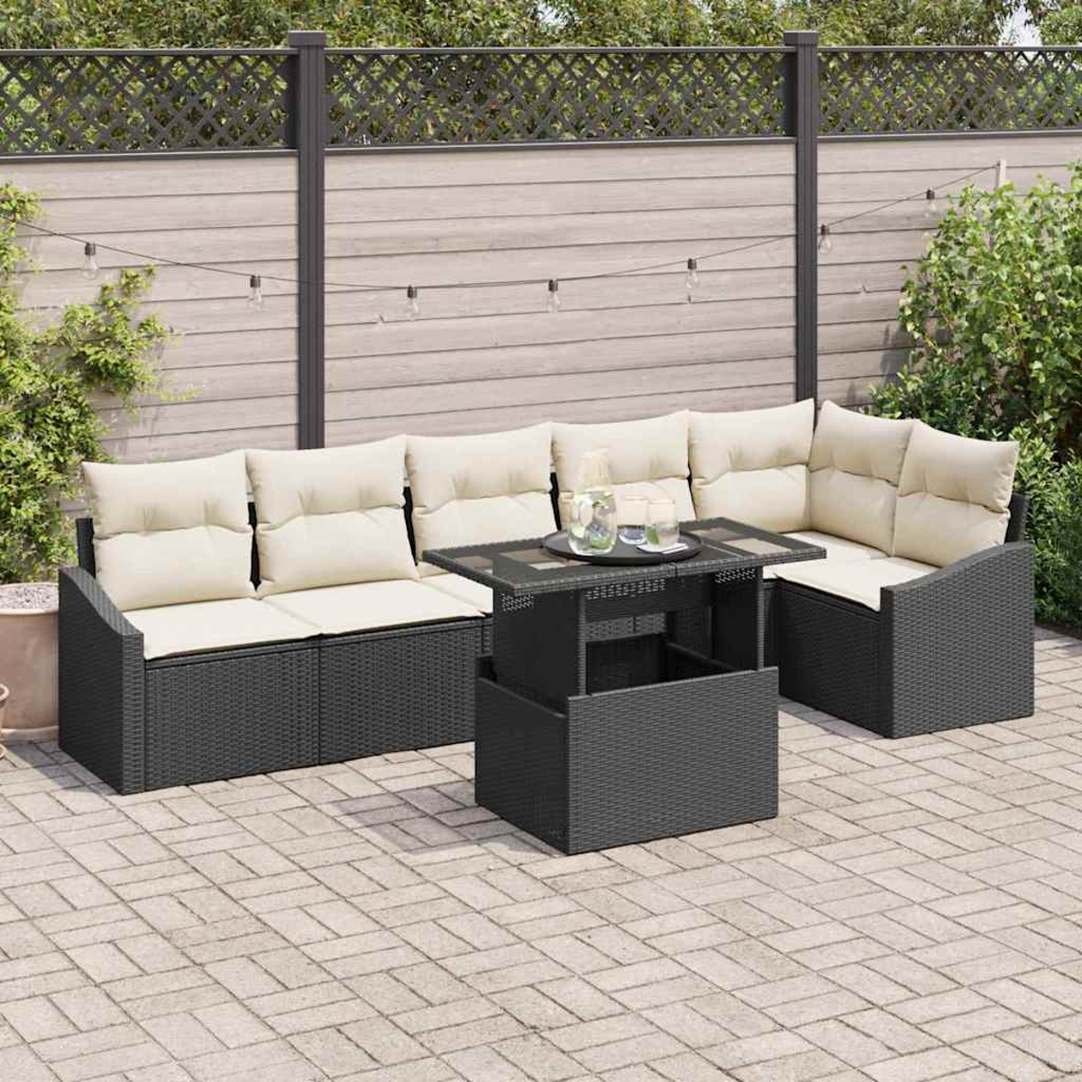 Juego de patio exterior: sofá de mimbre negro con cojines crema, una mesa a juego y un patio pavimentado. Una valla y vegetación al fondo.