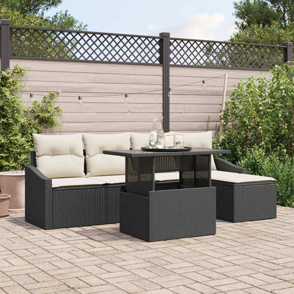 Garten-Terrassenset mit schwarzen Korbmöbeln, cremefarbenen Kissen und einem Tisch mit Hubfunktion. Im Hintergrund ein Holzzaun und Pflanzen.