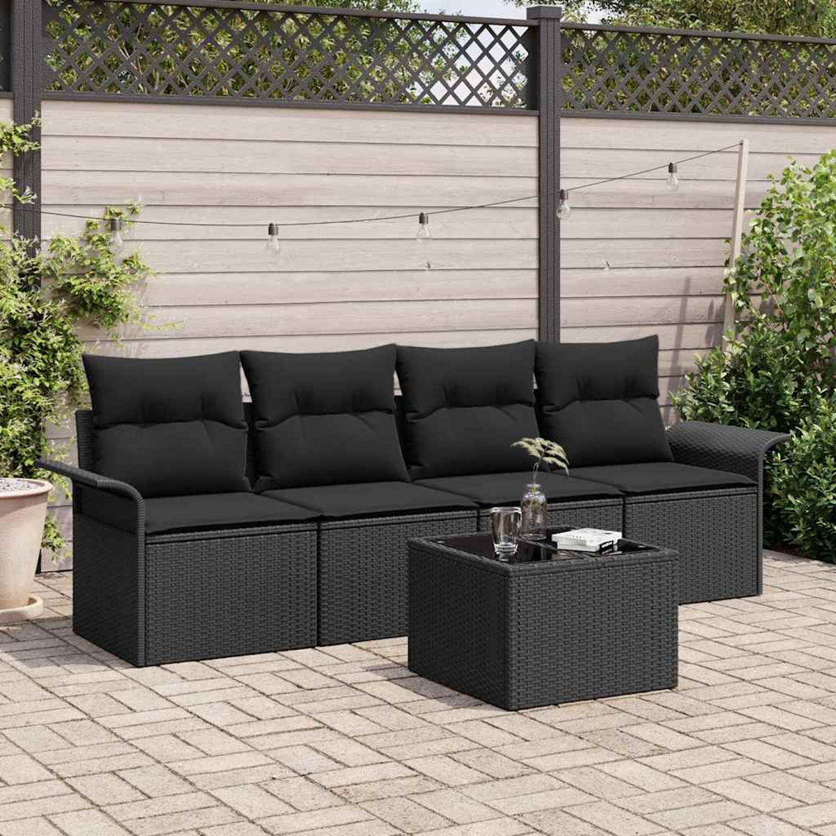 Ein schwarzes Korbmöbel mit schwarzen Kissen und ein passender Couchtisch auf einer Backstein-Terrasse. Ein Holzzaun und Pflanzen sind im Hintergrund.