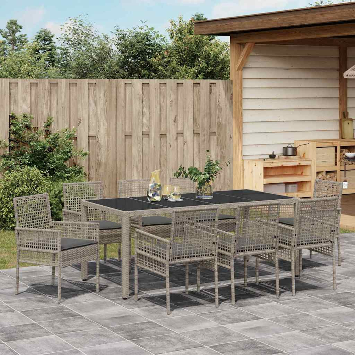 Essgarnitur im Freien auf einer Terrasse. Mit Tisch, Stühlen und einem Holzschuppen im Hintergrund. Grüne Pflanzen und Holzzaun sichtbar.