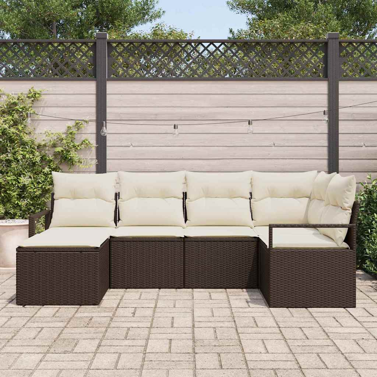 Ein braunes Outdoor-Sofa aus Korbgeflecht mit cremefarbenen Kissen steht auf einer Steinterrasse. Der Hintergrund umfasst einen Holzzaun und Pflanzen.