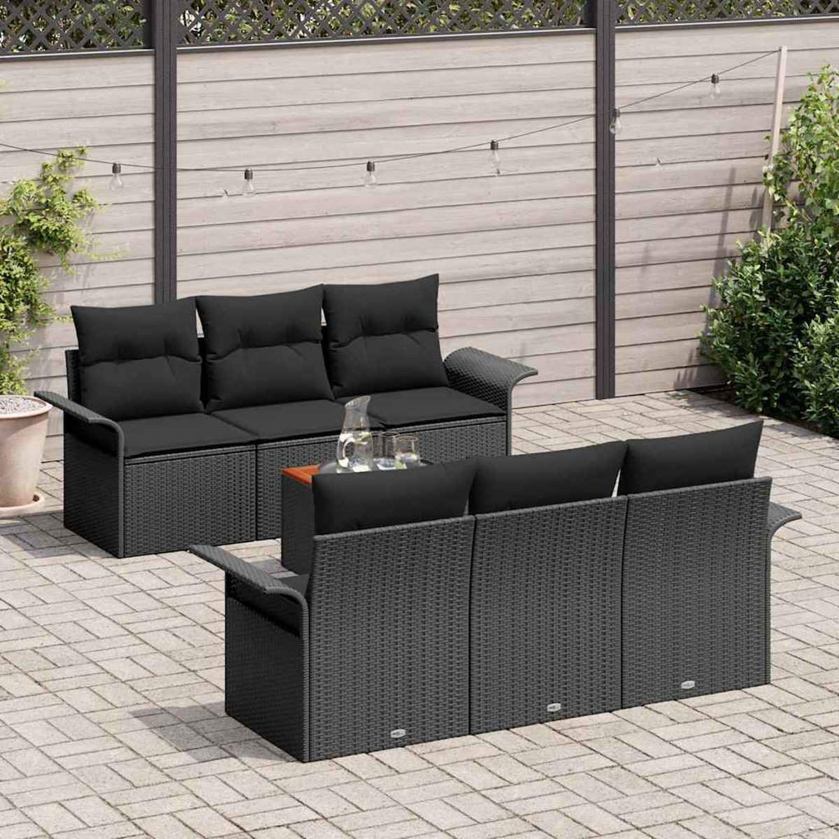 Szene im Freien mit zwei schwarzen Rattansofas, Kissen, einem kleinen Tisch mit Gläsern und einem Holzzaun mit Lichterketten.