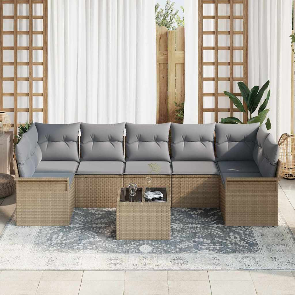Garten-Sitzgruppe mit grauen Kissen, Korbgeflecht-Basis und einem Tisch mit Glasplatte auf einem gemusterten Teppich. Weiße Vorhänge und Holzrahmen im Hintergrund.