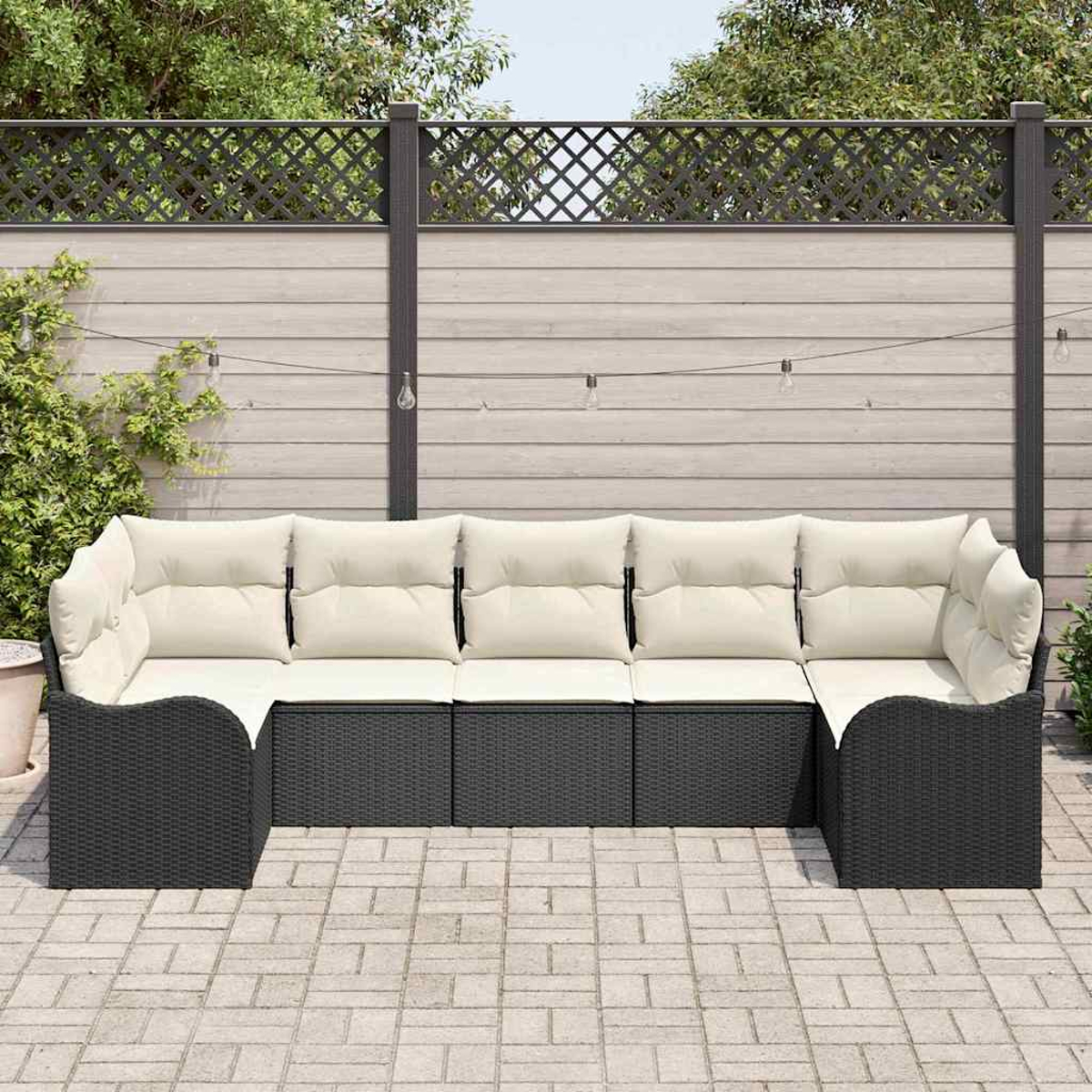 Ein schwarzes Korbsessel-Ecksofa mit cremefarbenen Kissen steht auf einer Backstein-Terrasse. Im Hintergrund ein Holzzaun und Grün.