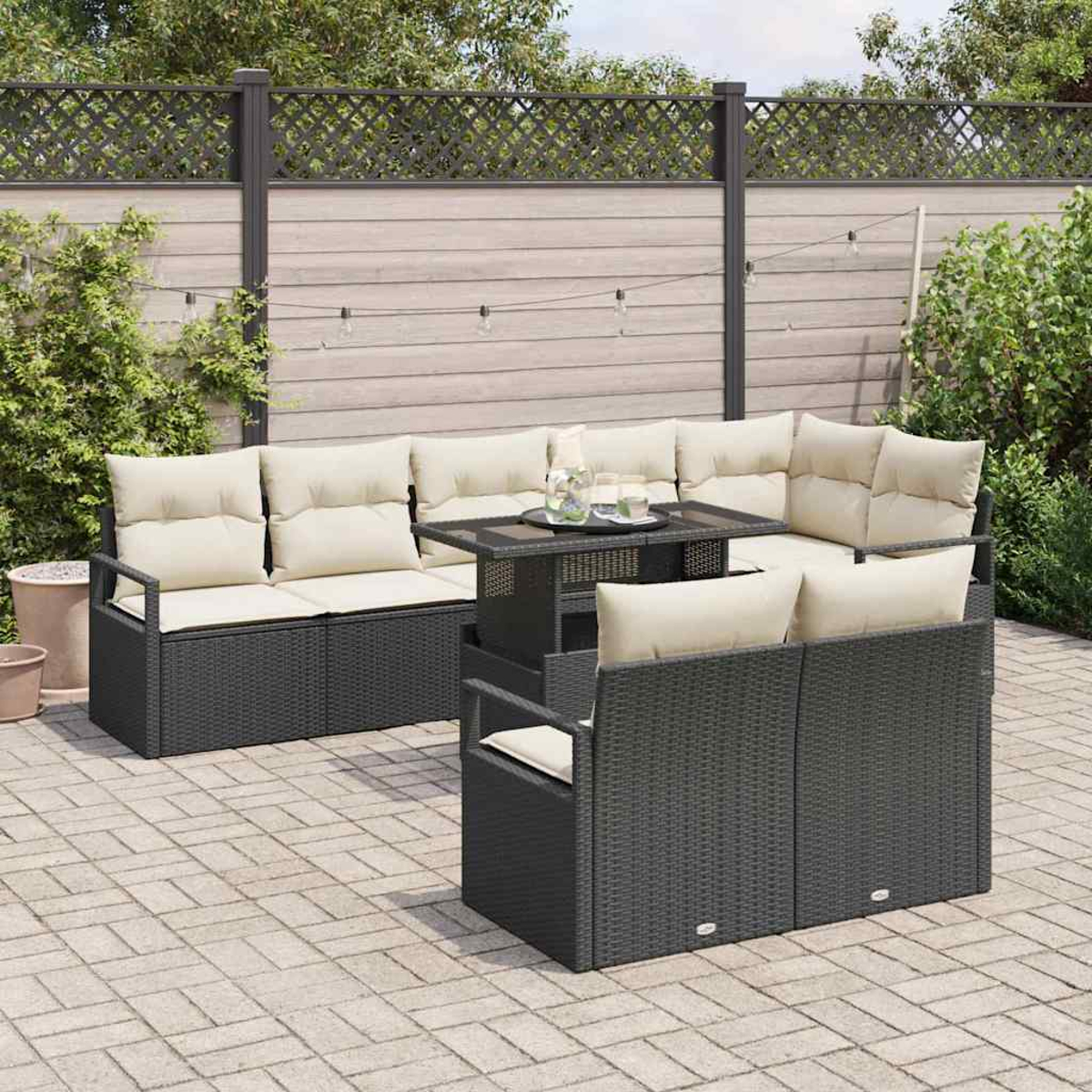 Garten-Terrassenset mit dunkelgrauen Korbmöbeln, cremefarbenen Kissen und einem Tisch mit Glasplatte. Vor Holzzaun und Grün.