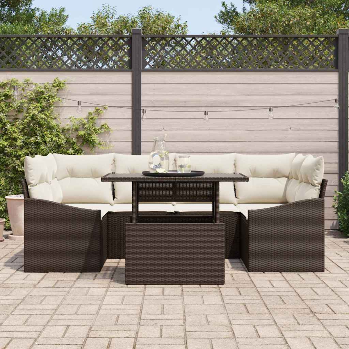 Terrassenset mit braunen Korbmöbeln, cremefarbenen Kissen und einem Tisch mit Glasplatte. Hintergrund: Holzzaun und Lichterketten.