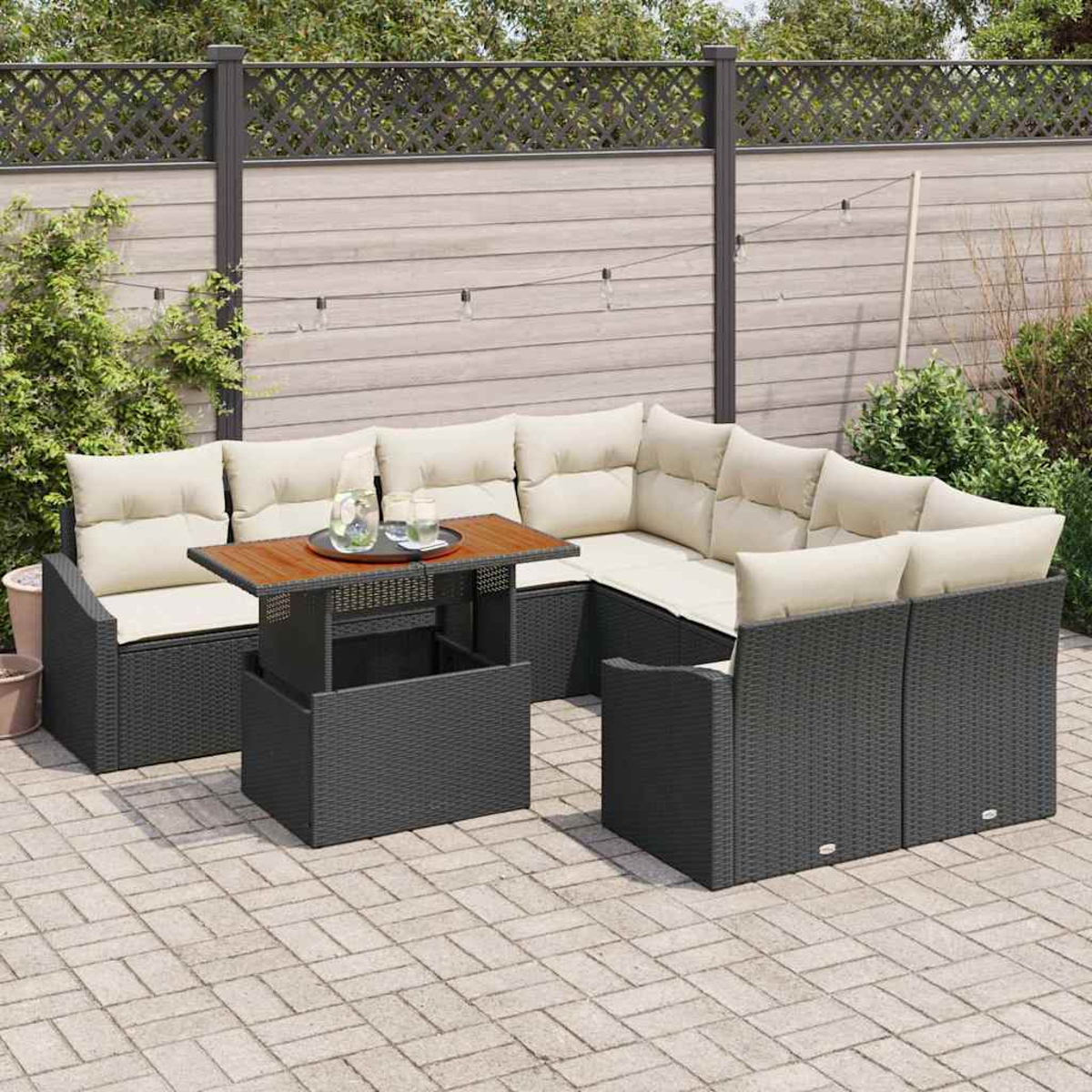 Garten-Terrassenset: graues Korbmöbel-Sofa mit cremefarbenen Kissen, Holztisch mit Getränken, auf Steinterrasse, Holzzaun im Hintergrund.