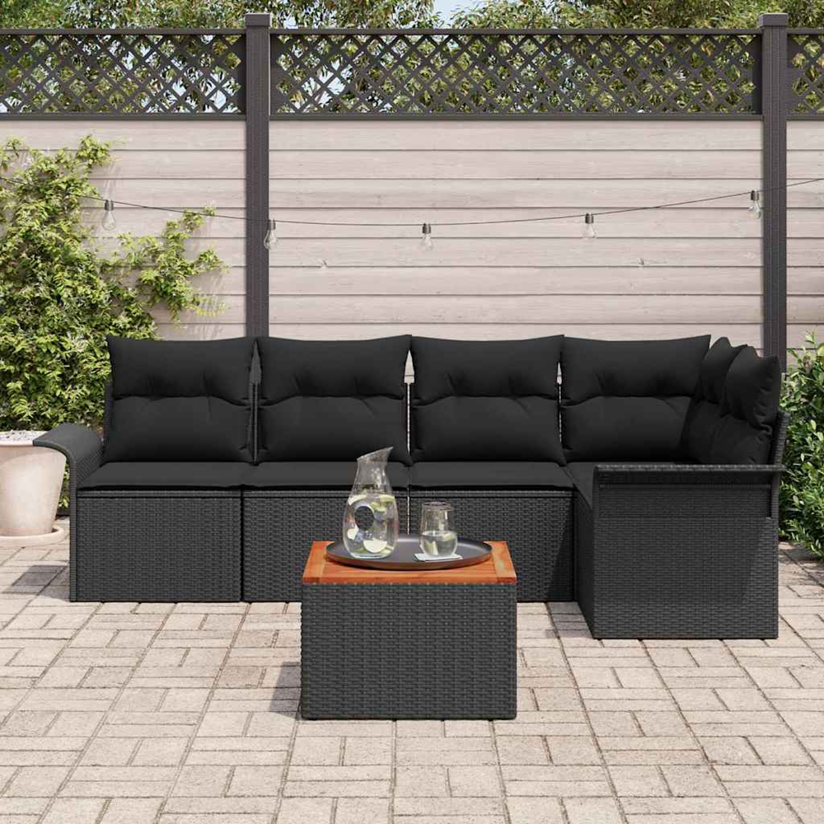 Terrassenset: schwarzes Korbmöbel mit schwarzen Kissen, Holztisch mit Tablett und Krug, auf Ziegelpflaster, mit Zaun.