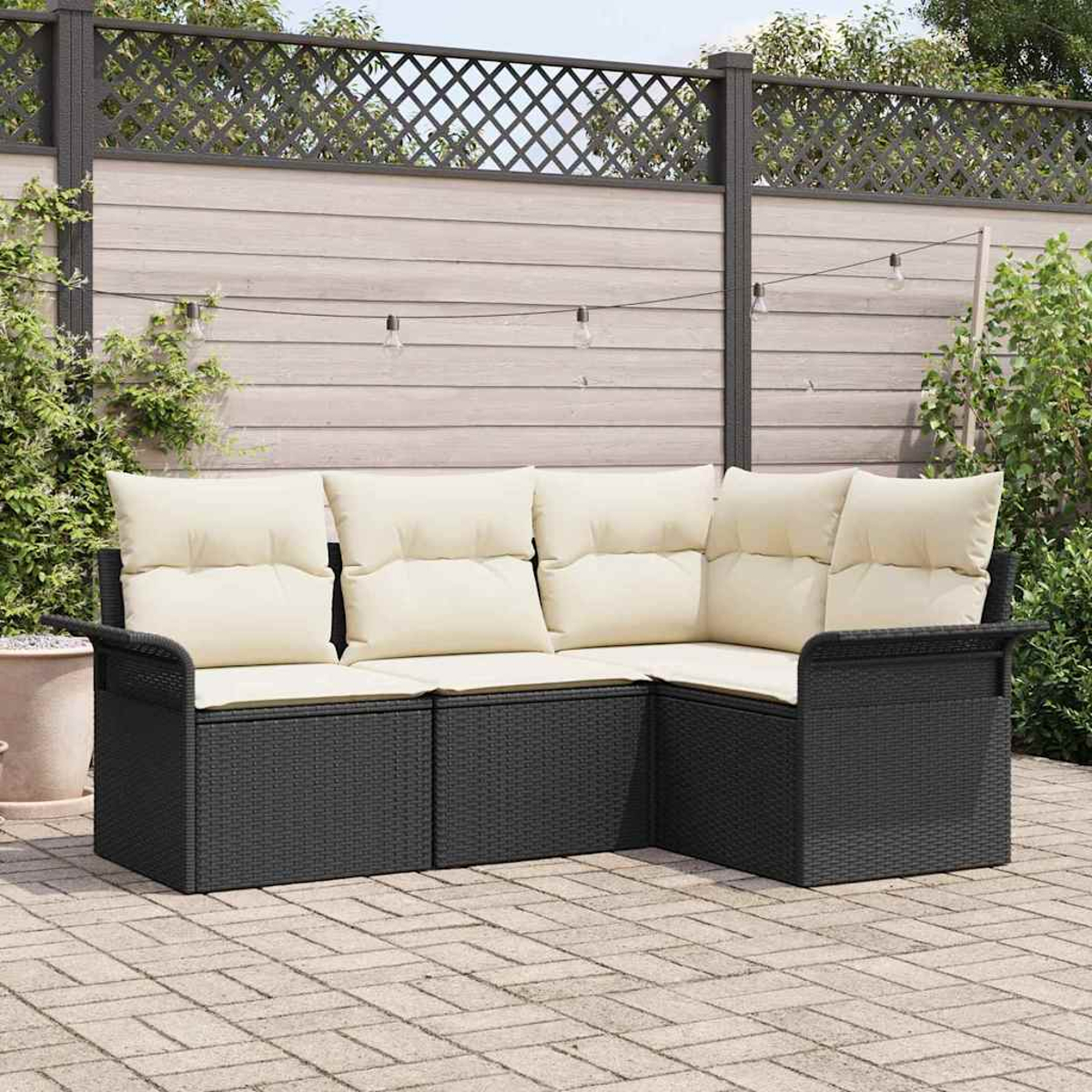 Schwarzes Outdoor-Geflecht-Ecksofa mit cremefarbenen Kissen auf einer Backstein-Terrasse. Holzzaun mit Lichterketten im Hintergrund.