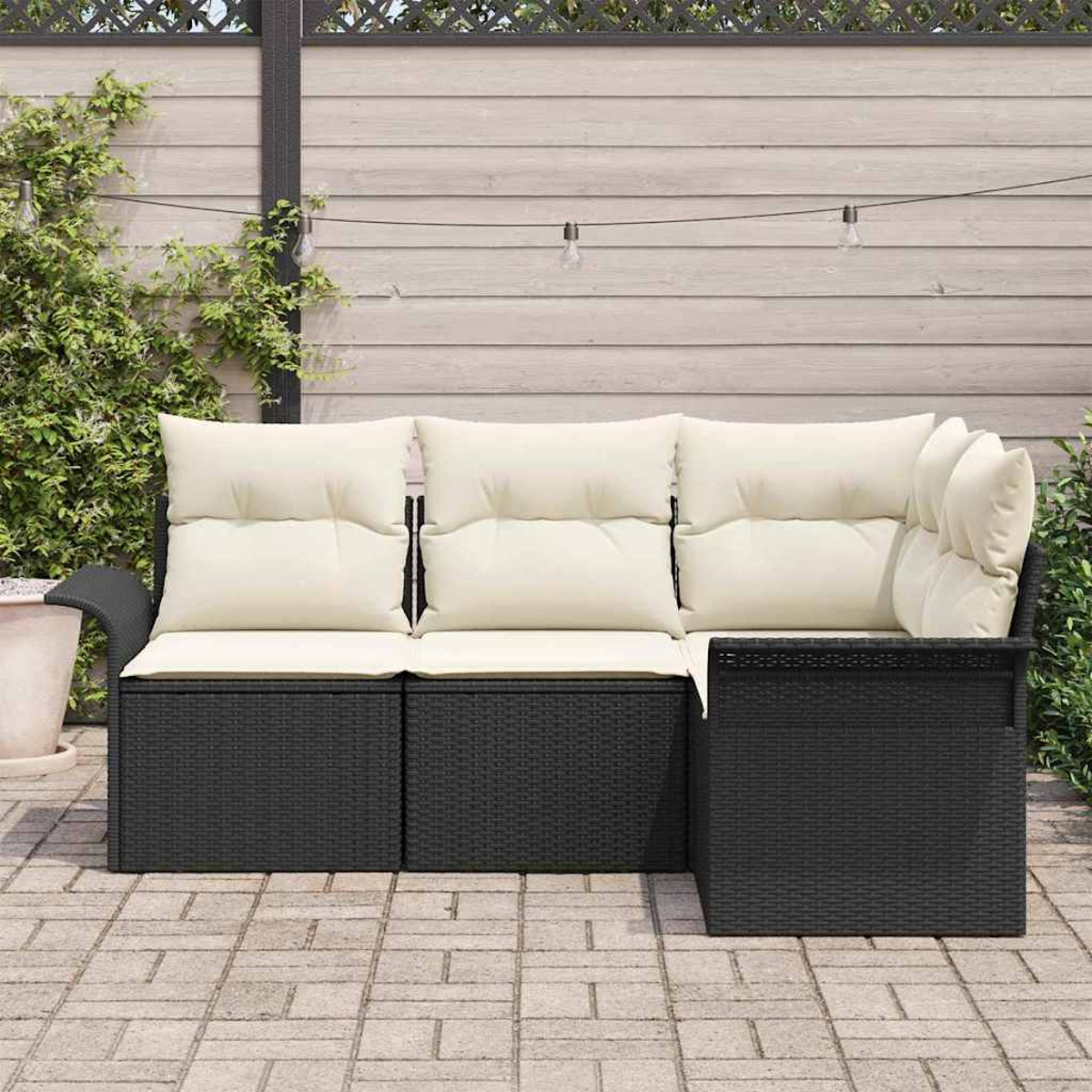Schwarzes Rattan-Outdoor-Sofa mit cremefarbenen Kissen steht auf einer Ziegelstein-Terrasse. Im Hintergrund ein Holzzaun und Grün.
