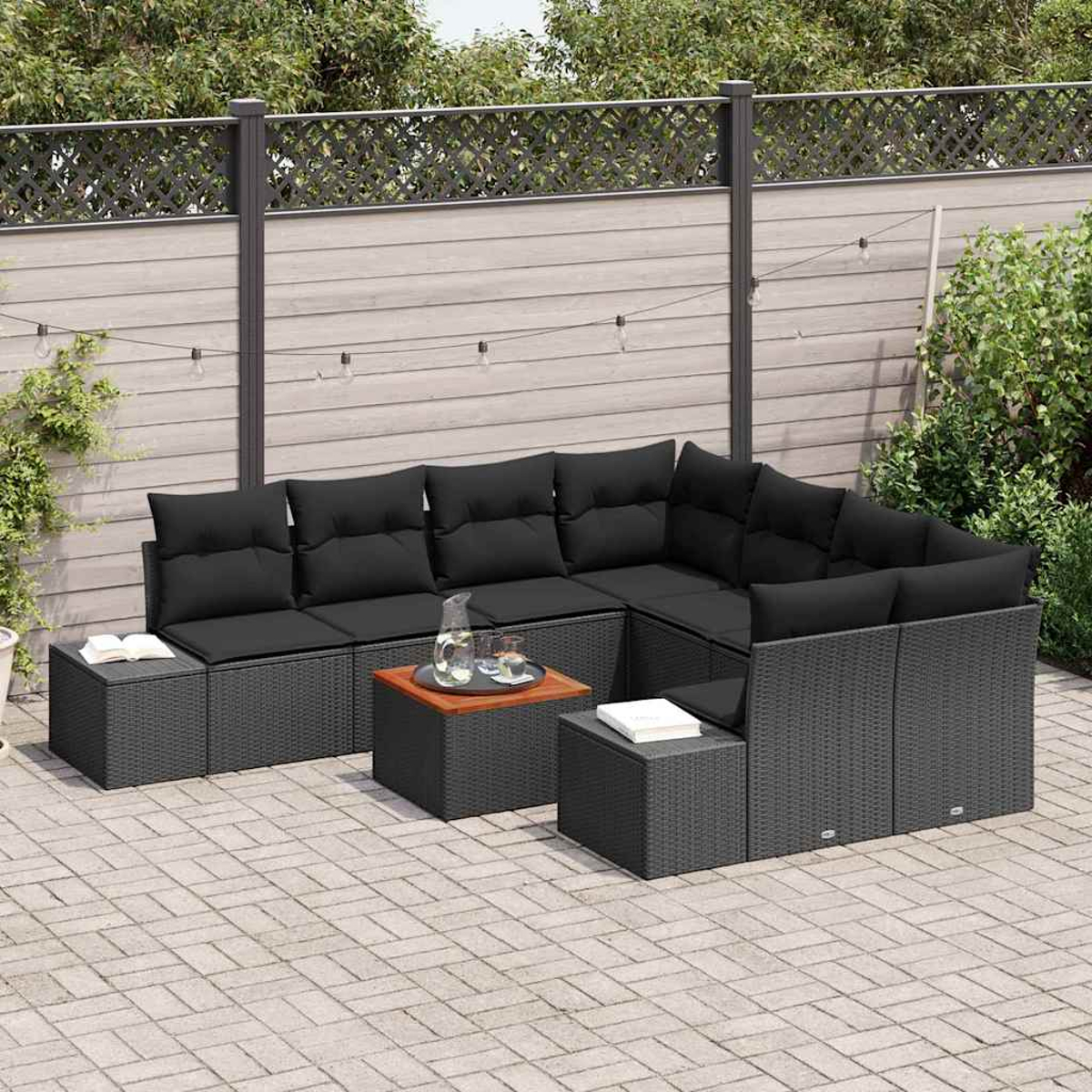 Outdoor-Ecksofa mit schwarzen Kissen und grauem Rattan. Zwei kleine Tische mit einem Buch und Glas. Hintergrund: Holzzaun und Pflanzen.