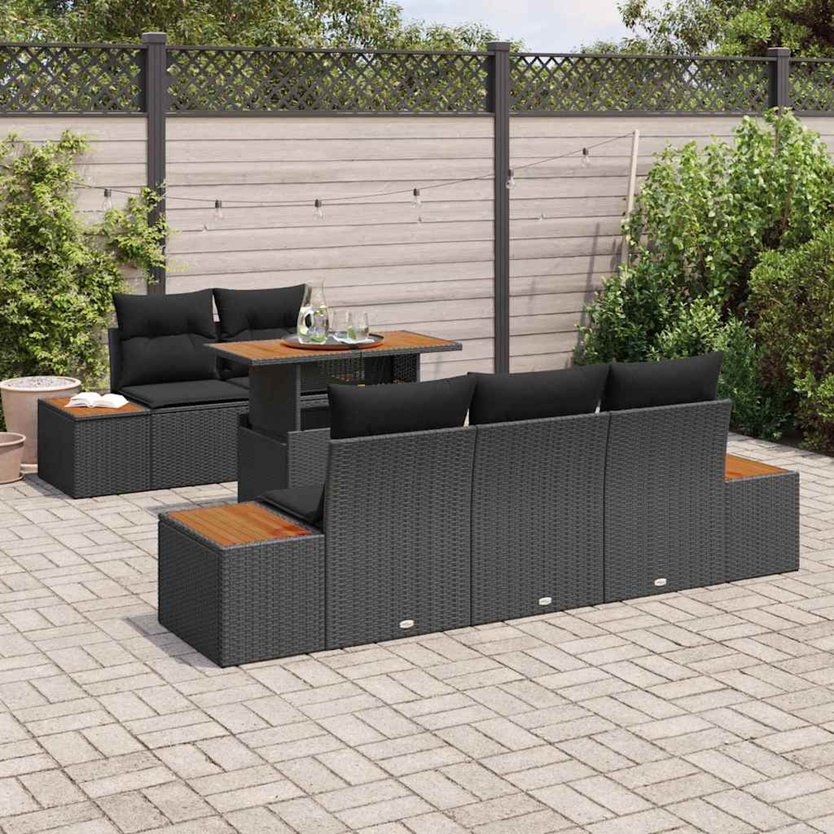 Outdoor-Terrassenset mit schwarzen Kissen und Holztisch. Graue Korbmöbel auf einer Ziegelterrasse, mit Zaun und Grün.