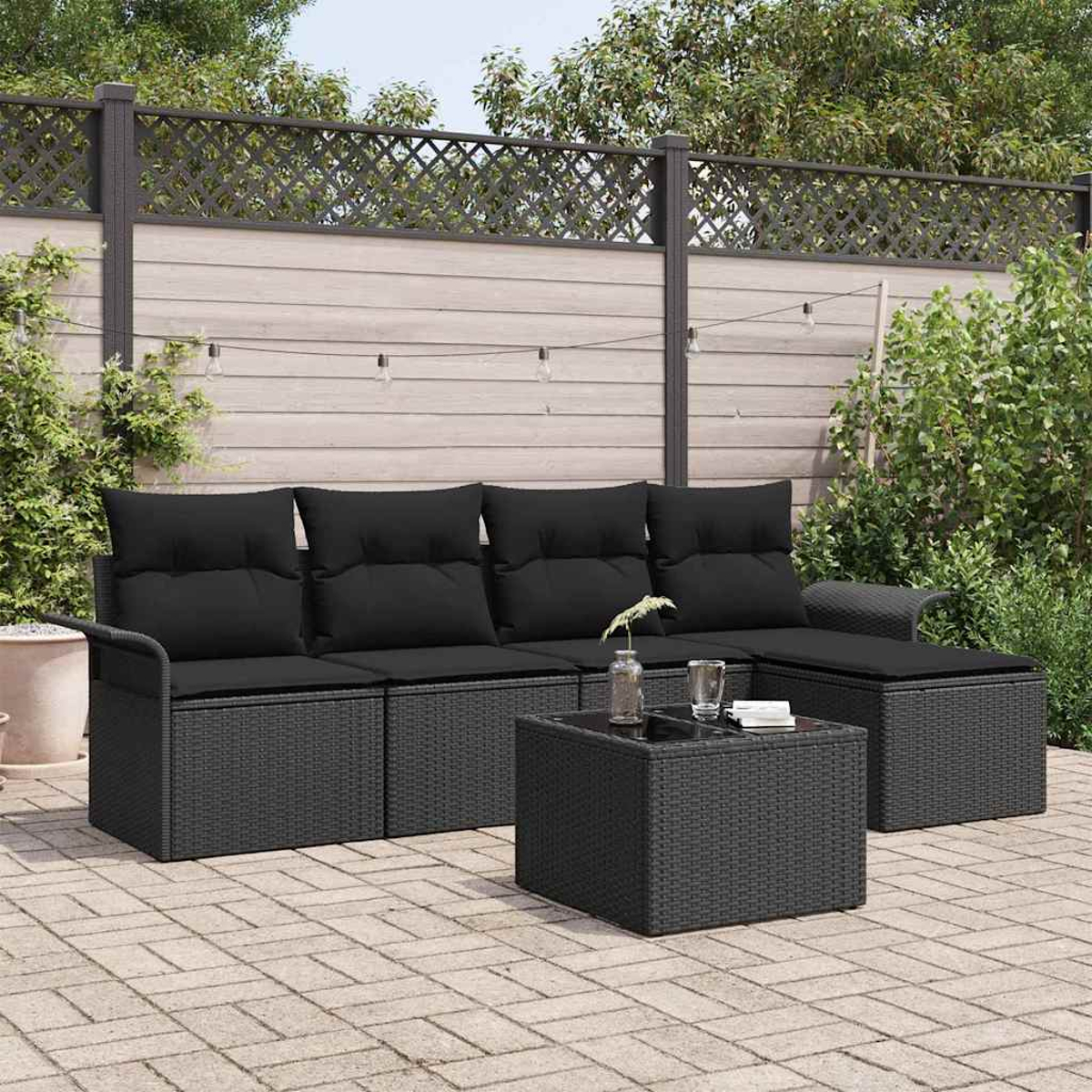 Garten-Terrassenset mit schwarzen Kissen und Tisch. Auf einer Backstein-Terrasse, umgeben von einem Zaun und Grün.