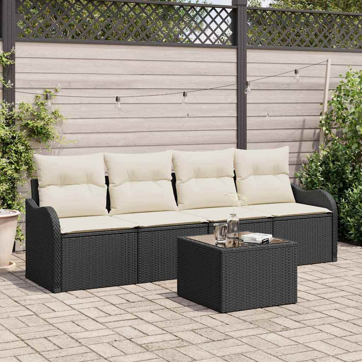 Garten-Terrassenset: schwarzes Korbmöbelsofa mit cremefarbenen Kissen, passender Tisch, Lichterkette und Holzzaun. Gepflasterte Terrasse mit Grün.