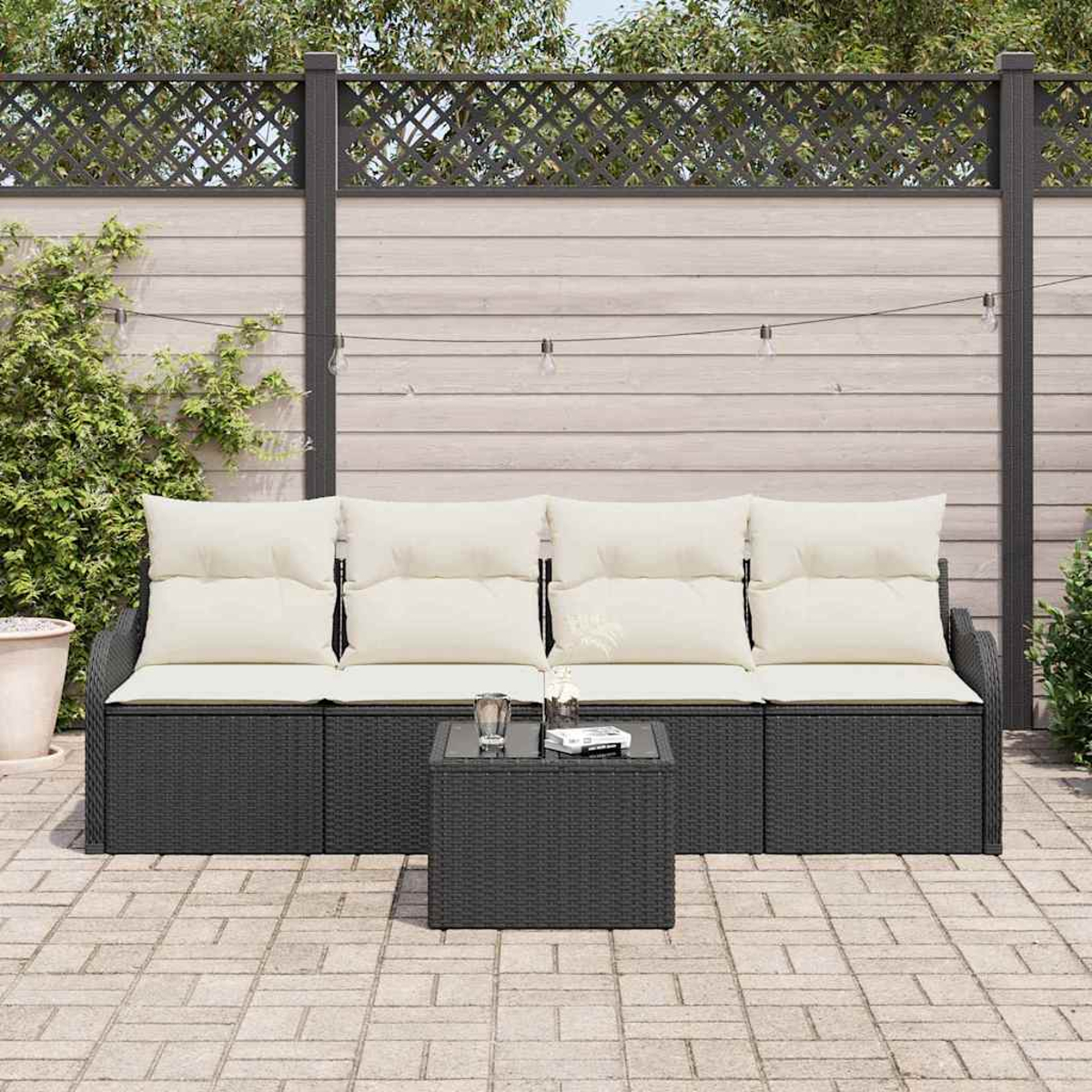 Dunkelgraues Rattan-Sofa mit cremefarbenen Kissen und einem kleinen Tisch auf einer Terrasse. Dahinter ein Holzzaun und Grün.