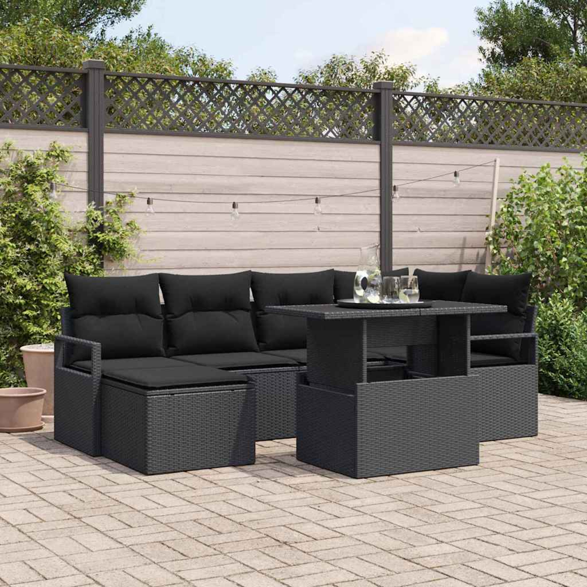 Garten-Terrassenset: schwarzes Korbmöbel-Sofa, Tisch und Kissen auf einer Backstein-Terrasse, mit Holzzaun und Grün im Hintergrund.