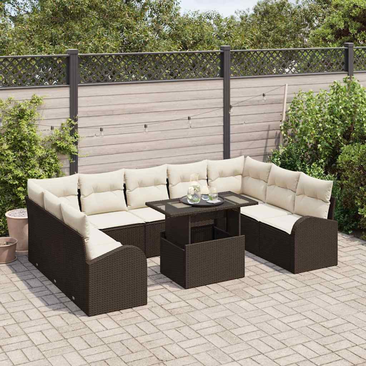 Gartenmöbel-Set mit braunem Korbgeflecht und cremefarbenen Kissen, Tisch mit Gläsern. Hintergrund mit Zaun und Grün.