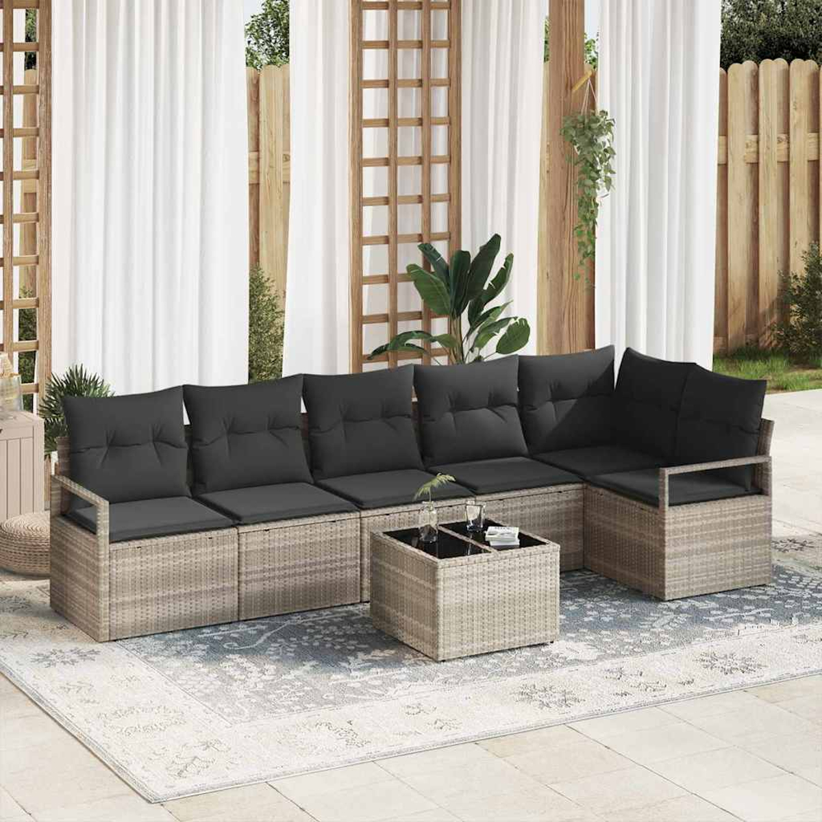 Gartensitzgruppe mit grauen Kissen, Couchtisch und Teppich. Weiße Vorhänge und Holzgitter im Hintergrund, Pflanzen vorhanden.