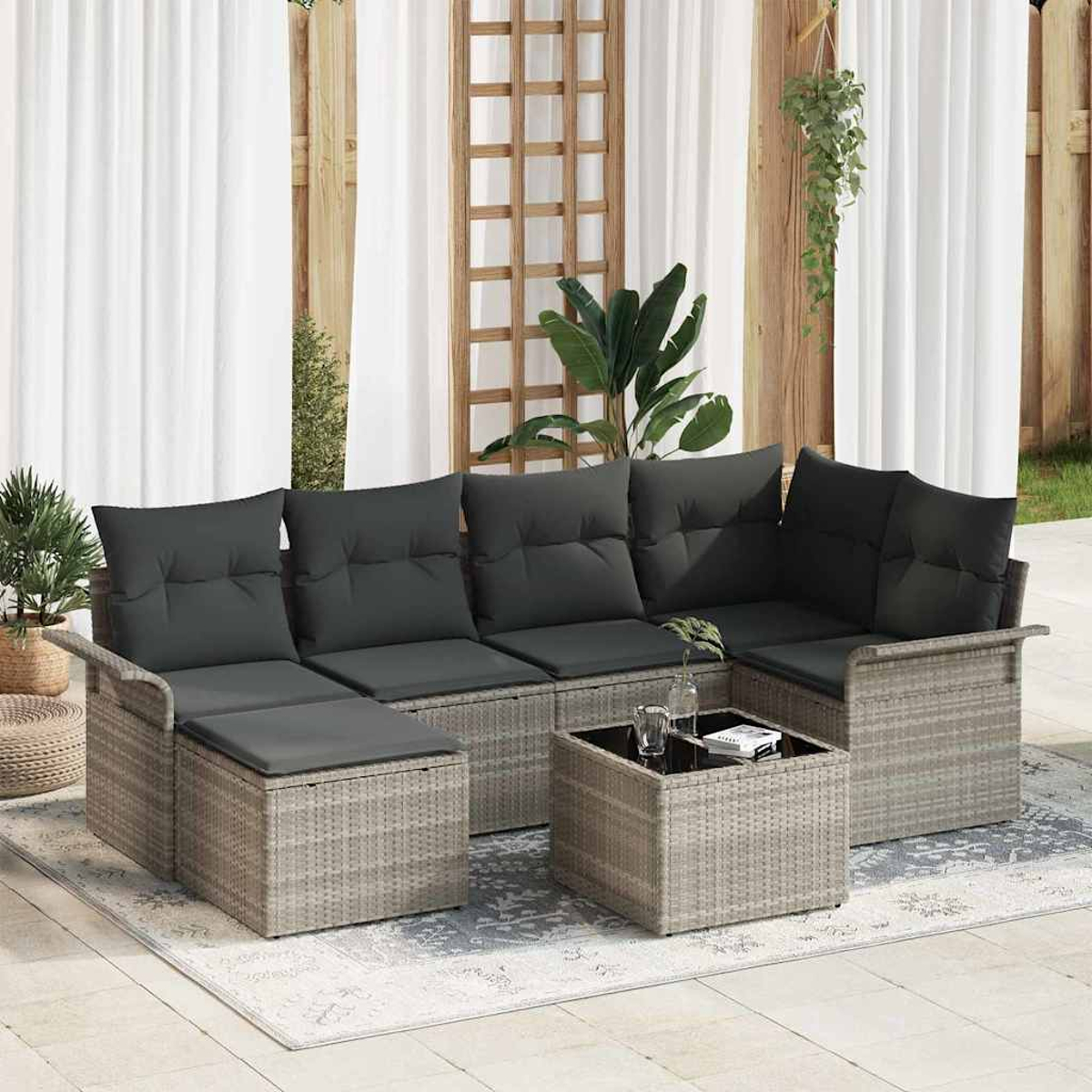 Outdoor-Korbsessel mit grauen Kissen, Couchtisch mit Glasplatte und Pflanzen. Weiße Vorhänge und Holzgitter im Hintergrund.
