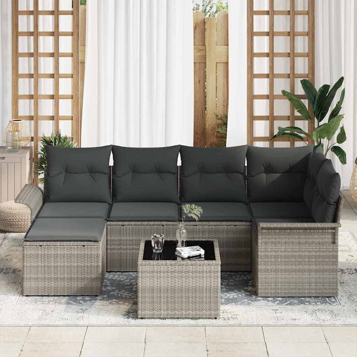 Gartensofa mit dunklen Kissen, Tisch mit Glasplatte und Teppich. Vorhänge und Holzgitter im Hintergrund.