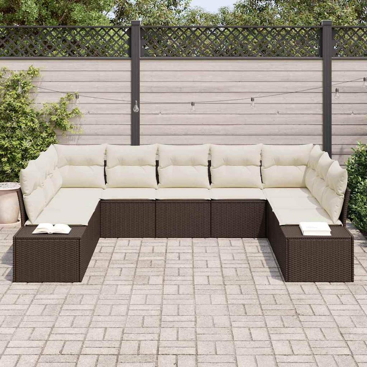 Ein U-förmiges braunes Korbmöbel-Sofa mit beigen Kissen steht auf einer Terrasse. Im Hintergrund befinden sich ein Zaun und Pflanzen.
