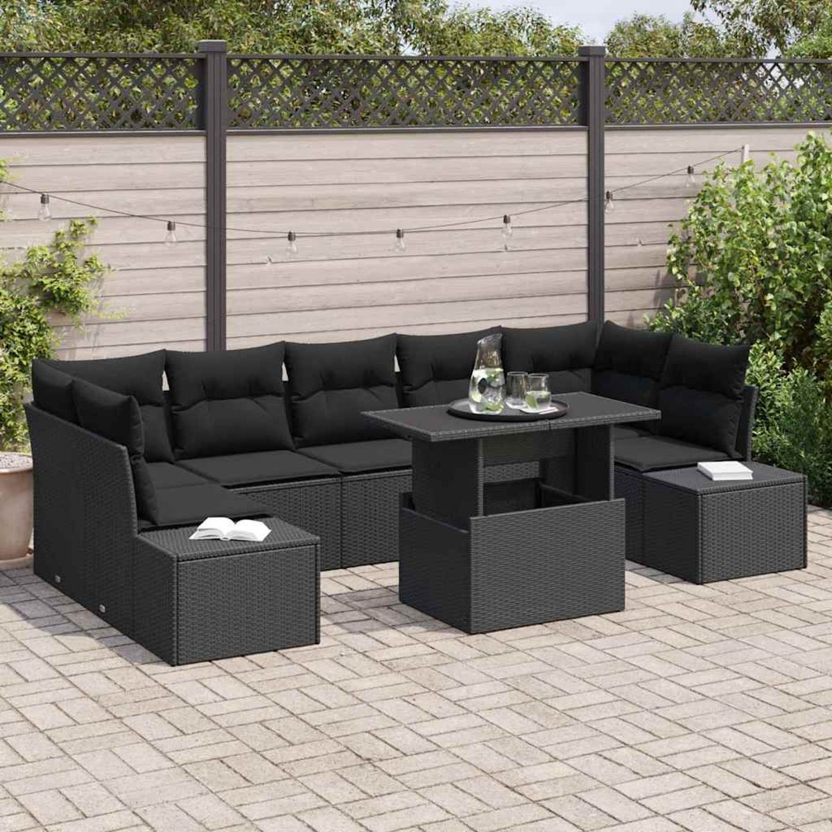 Schwarzes Outdoor-Ecksofa aus Korbgeflecht mit Kissen, Tisch und Hocker. Im Hintergrund ein Zaun und Grün. Ein Buch liegt auf dem Hocker.