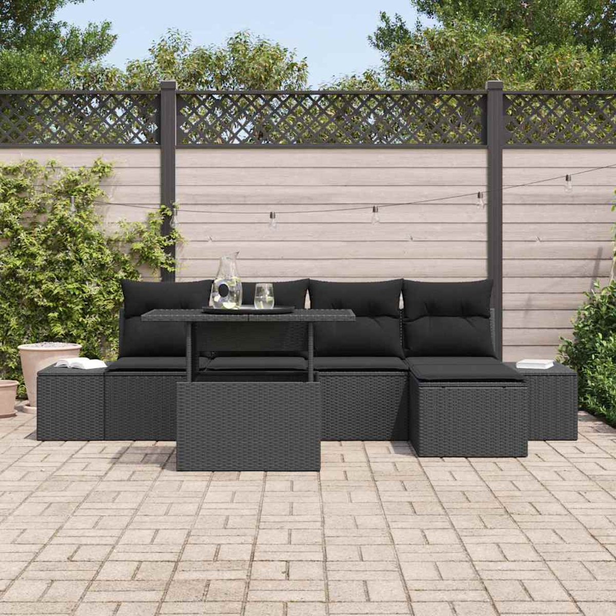 Terrassenset mit schwarzen Kissen. Enthält Tisch, Sofa und Hocker. Vor Holzzaun mit Grün.