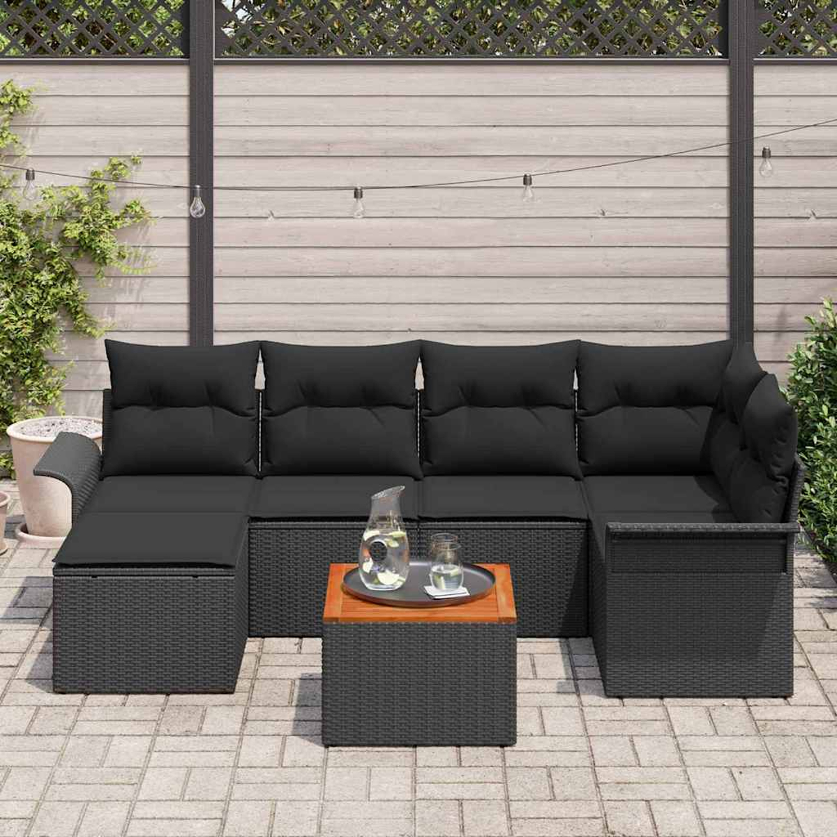 Outdoor-Ecksofa mit schwarzen Kissen, Holztisch mit Glaskrug und Glas. Auf einer Terrasse aufgestellt.