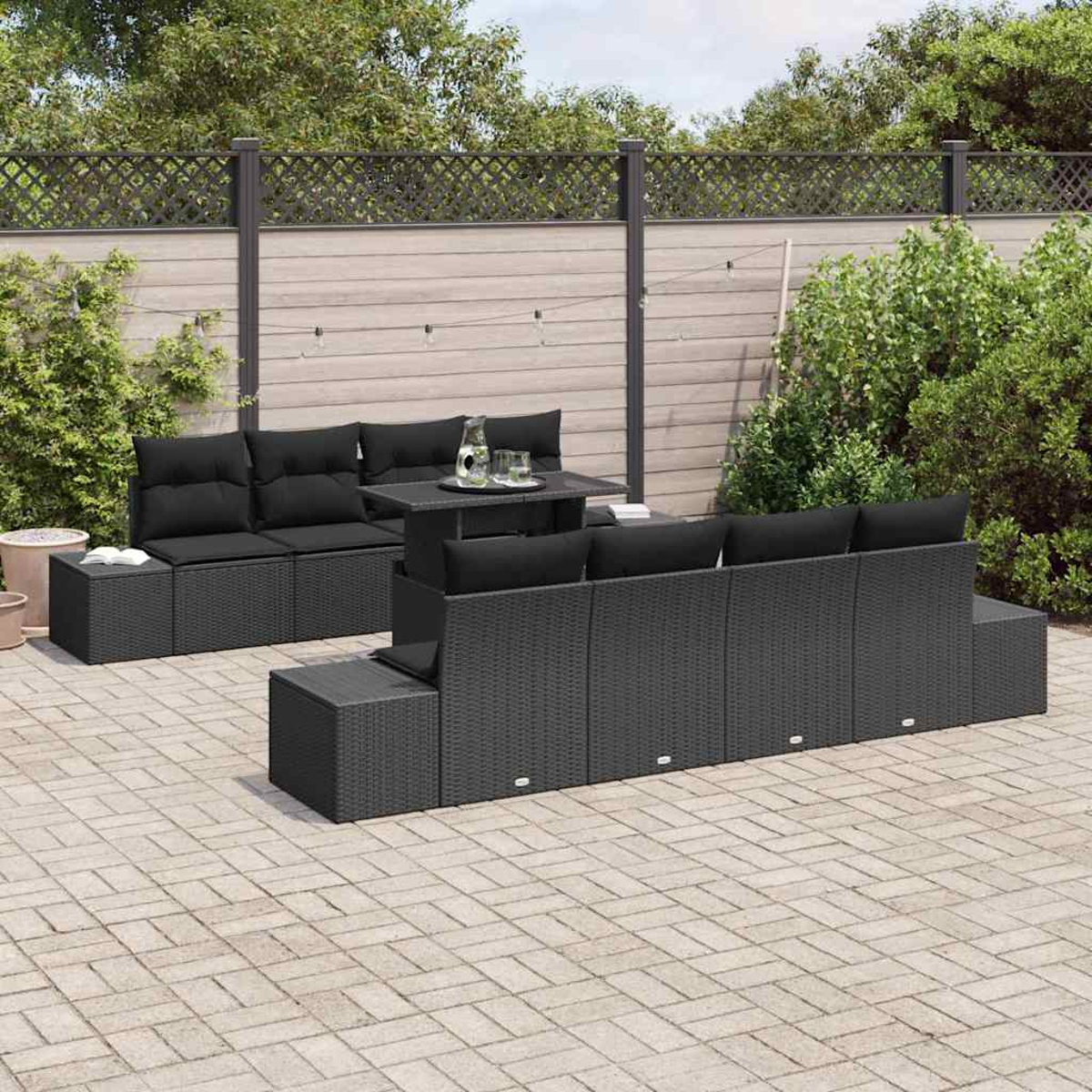 Escena al aire libre con juego de muebles de mimbre negro. Incluye sofás, mesa y otomanas en un patio de ladrillos, rodeado de vegetación y una valla.