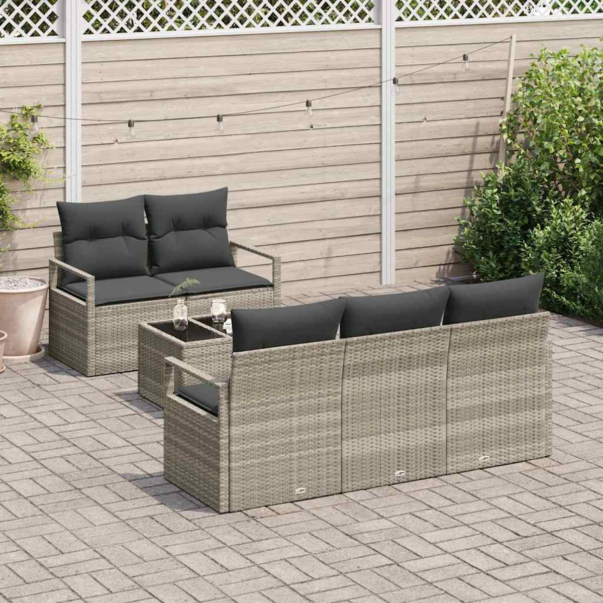 Garten-Rattan-Möbel-Set mit grauen Kissen auf einer gepflasterten Terrasse. Zwei Sofas und ein Tisch sind sichtbar, vor einem Holzzaun.