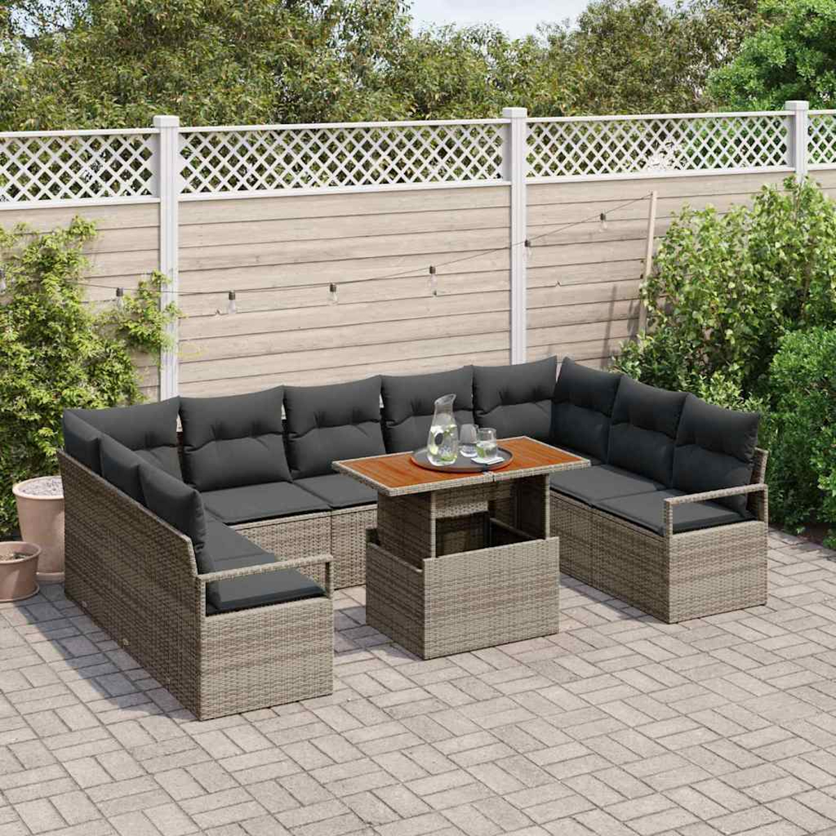 Geflochtenes Outdoor-Sofaset mit dunkelgrauen Kissen und einem Holztisch. Ein weißer Zaun und Grünzeug im Hintergrund.