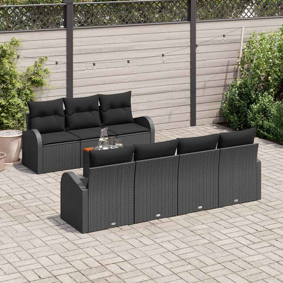 Gartenmöbel-Set mit schwarzen Kissen. Das Set umfasst ein Sofa und mehrere Sessel auf einer gepflasterten Terrasse. Im Hintergrund ein Holzzaun.