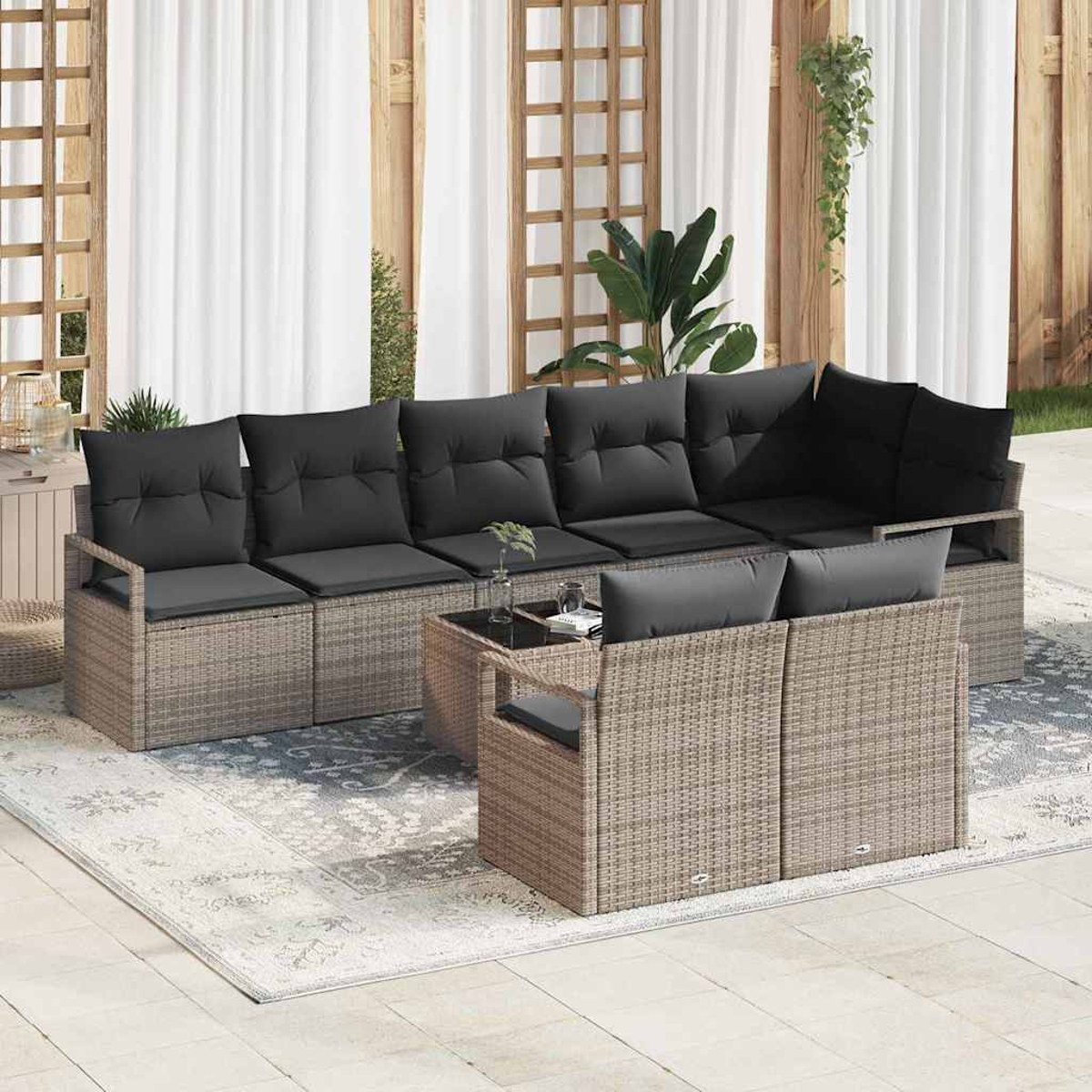 Rattan-Sofa im Freien mit schwarzen Kissen und einem Tisch mit Glasplatte. Vorhänge, Holzverkleidung und ein gemusterter Teppich vervollständigen das Bild.