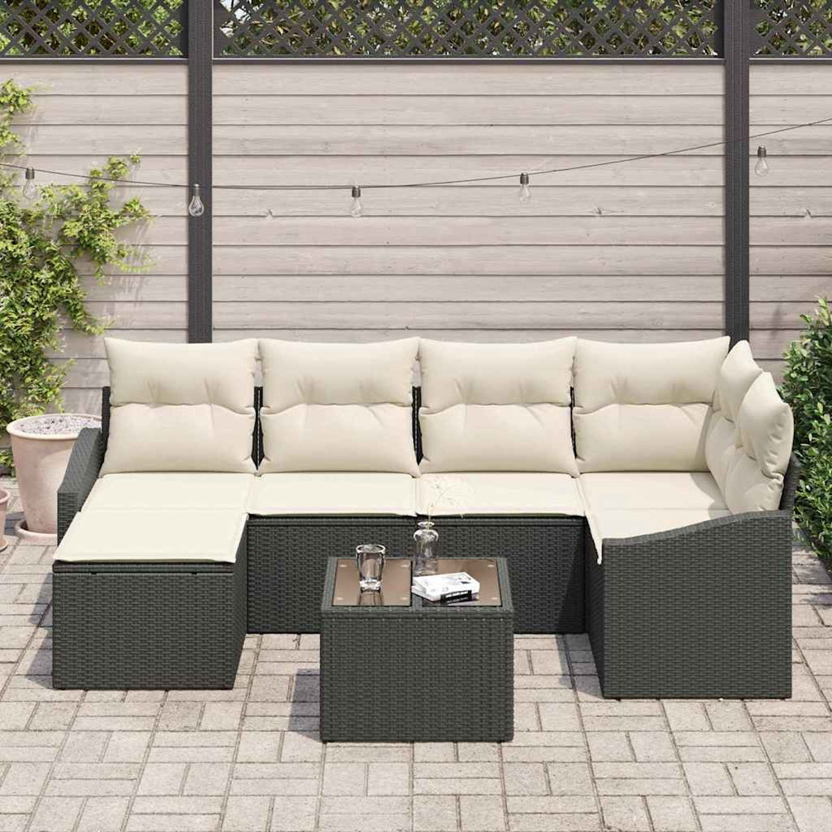 Juego de muebles de patio. Sofá de mimbre gris oscuro con cojines crema, mesa pequeña y valla de madera al fondo.
