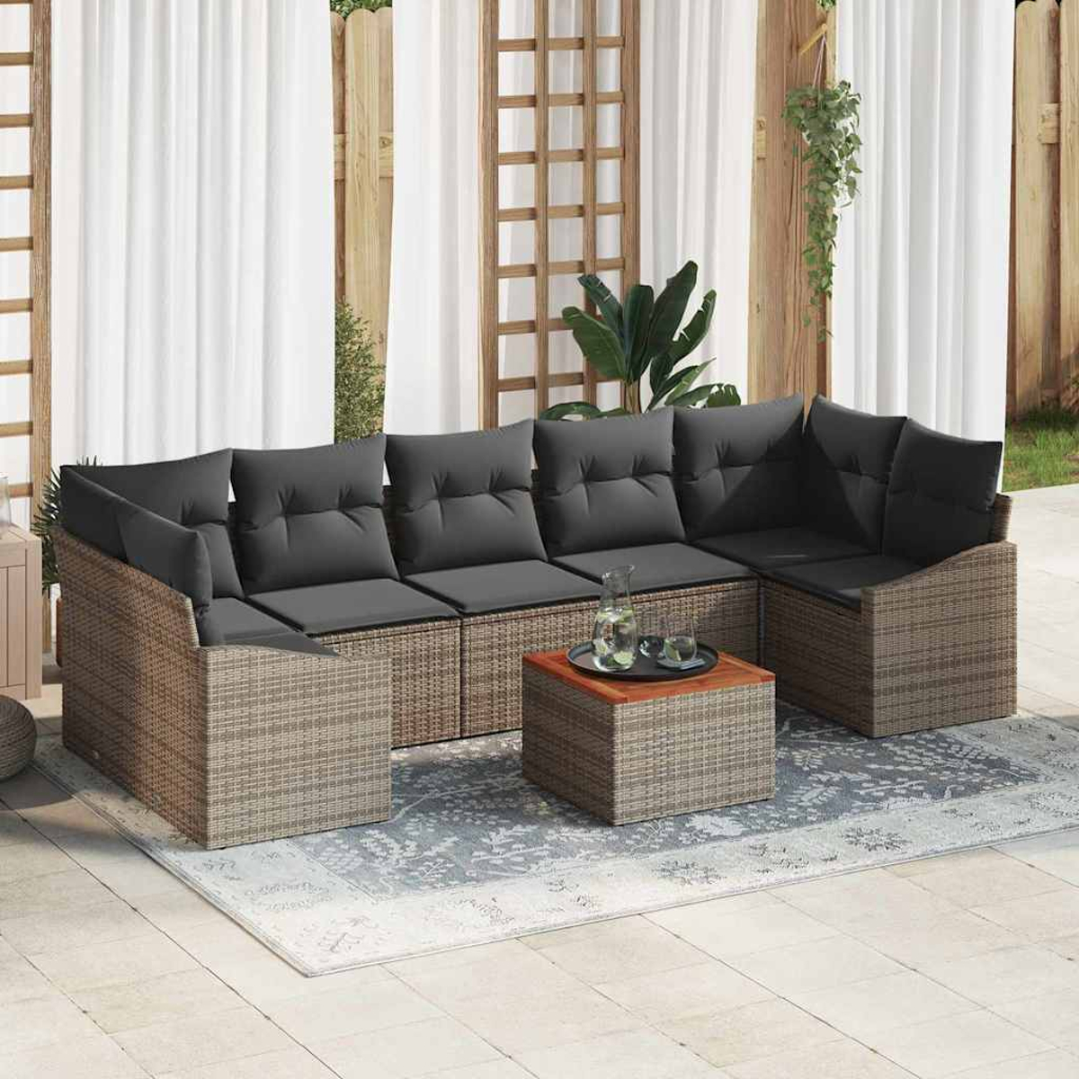 Gartenlounge mit grauen Kissen auf Korbmöbeln, kleinem Tisch und gemustertem Teppich. Weiße Vorhänge und Holzzaun.