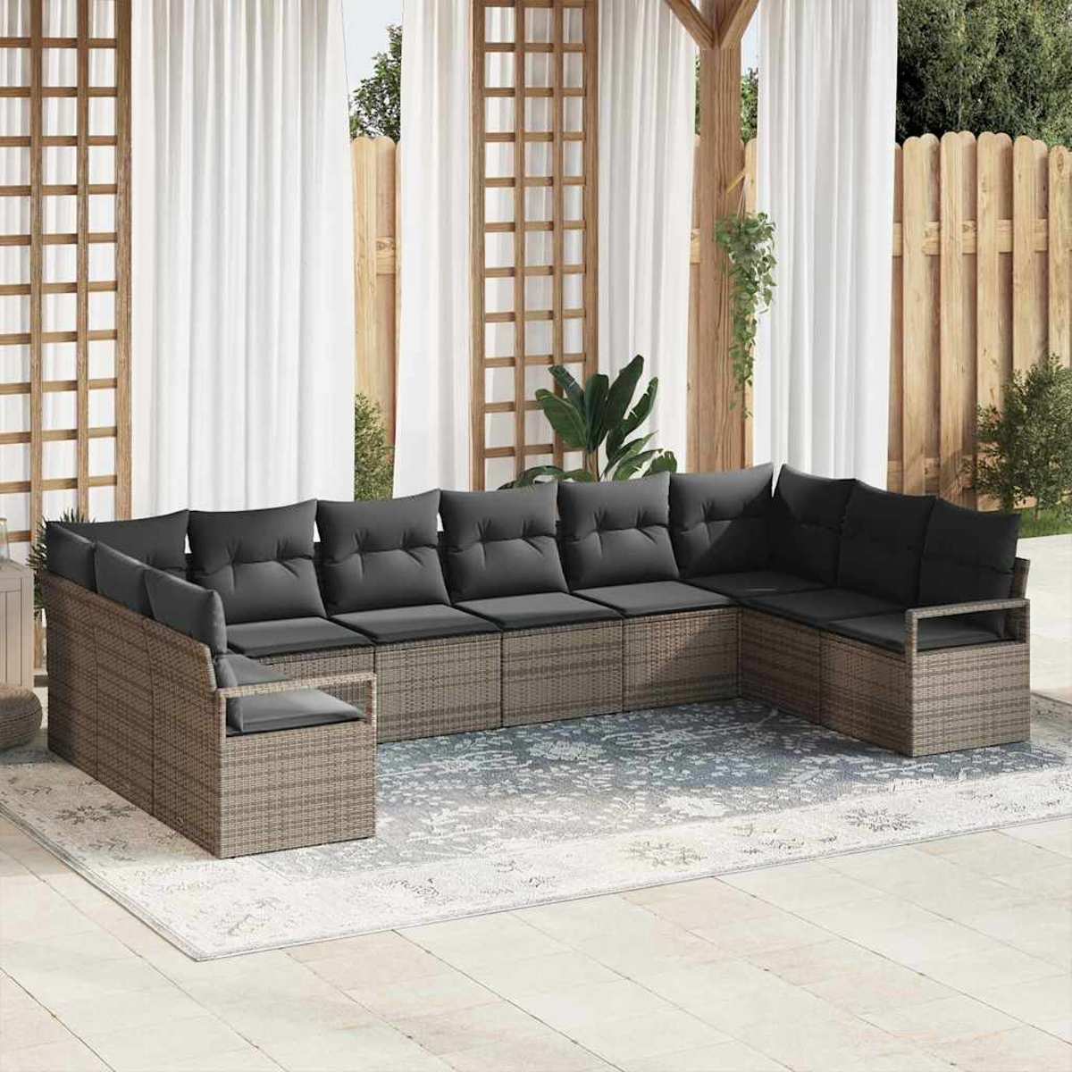 Ein Ecksofa im Freien mit grauen Kissen steht auf einem gemusterten Teppich. Weiße Vorhänge und ein Holzzaun bilden den Hintergrund.