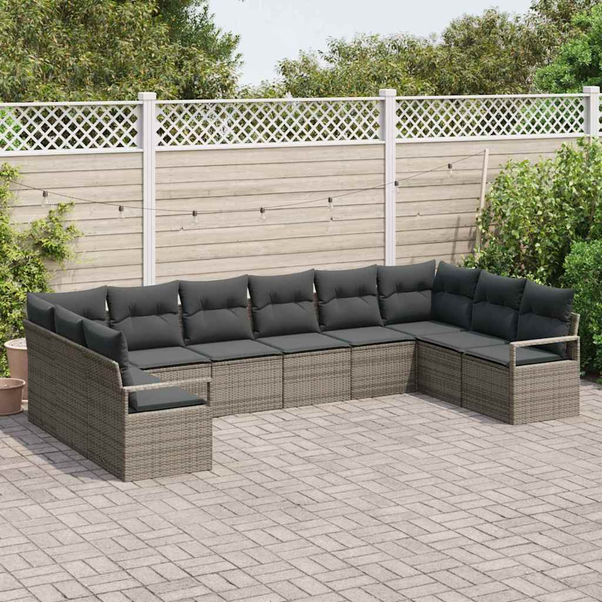 U-förmiges graues Outdoor-Sofa aus Korbgeflecht mit dunkelgrauen Kissen auf einer gepflasterten Terrasse. Holzzaun und Grün im Hintergrund.