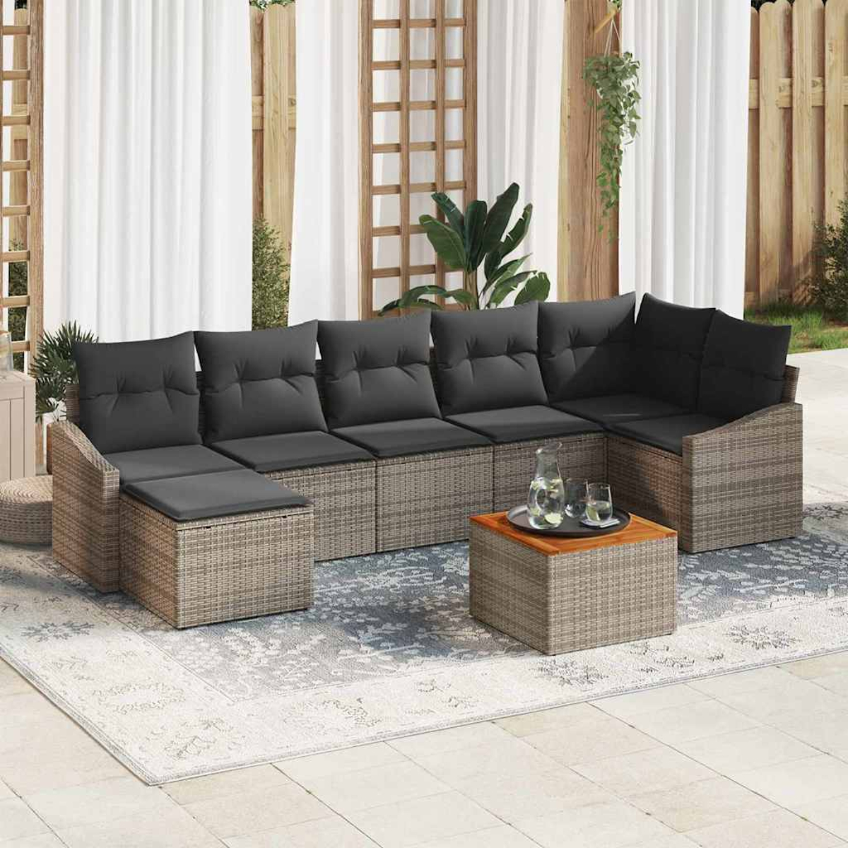 Ein graues Korbmöbel-Ecksofa mit schwarzen Kissen und ein Holztisch stehen im Freien auf einem gemusterten Teppich. Vorhänge und Pflanzen sind sichtbar.