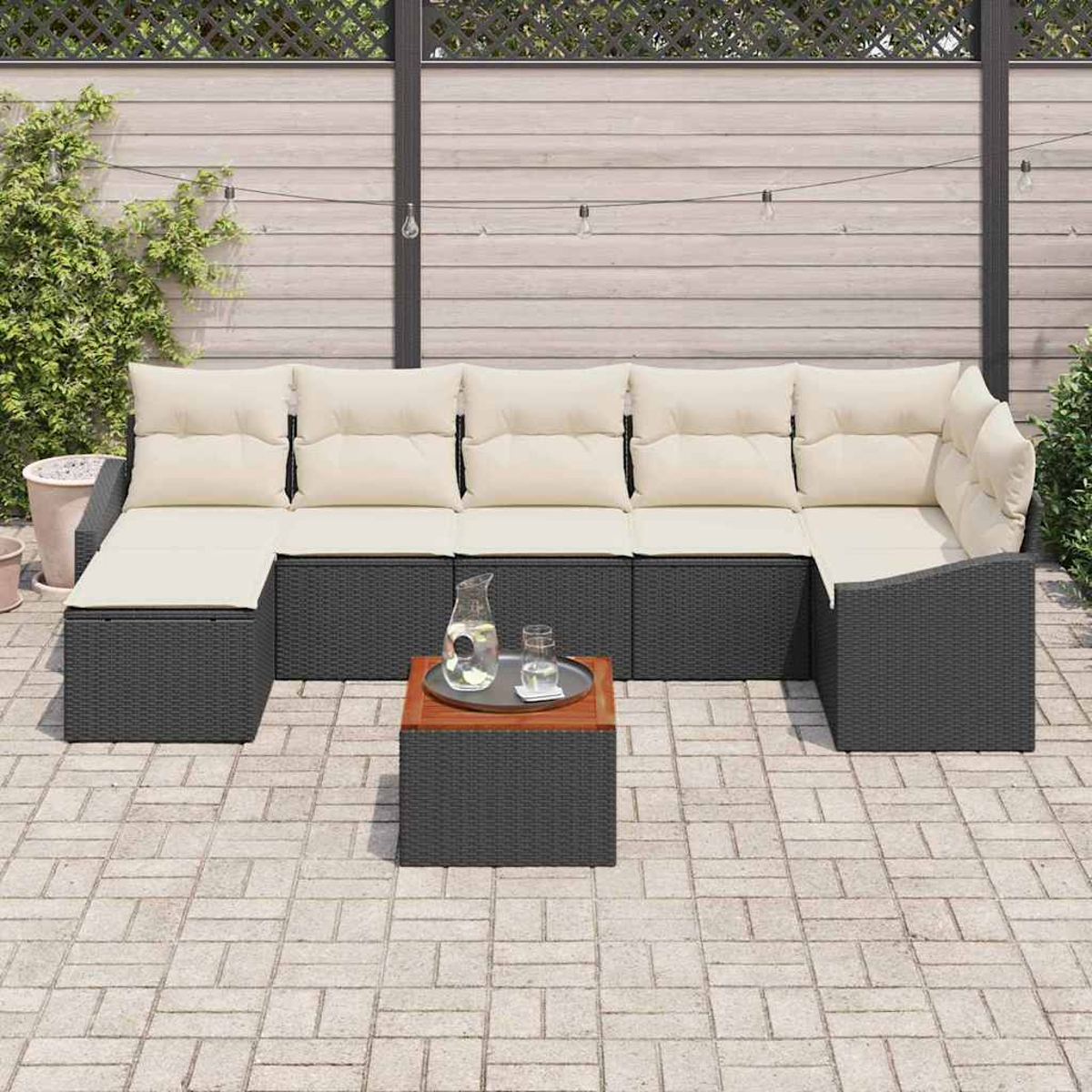Outdoor-Ecksofa mit cremefarbenen Kissen und dunkelgrauem Rahmen. Kleiner Tisch mit Krug, Gläsern. Ziegelpflaster mit Holzzaun im Hintergrund.