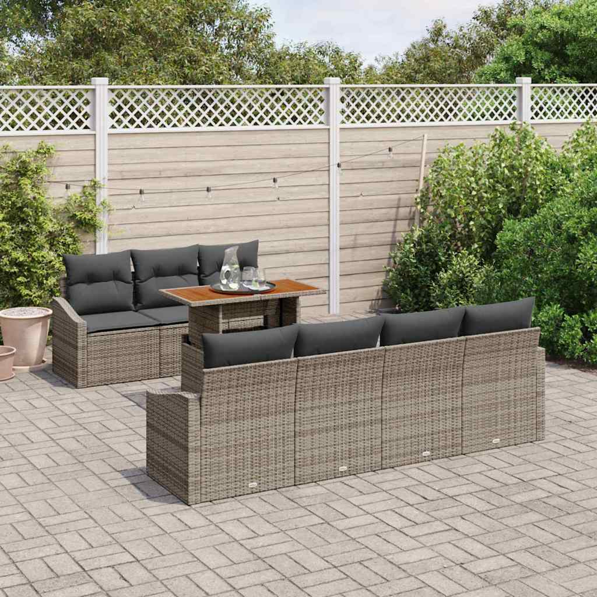 Szene im Freien mit Rattan-Sofagarnituren und einem Tisch. Im Hintergrund befindet sich ein weißer Zaun und grüne Büsche. Auf dem Tisch stehen ein Krug und Gläser.